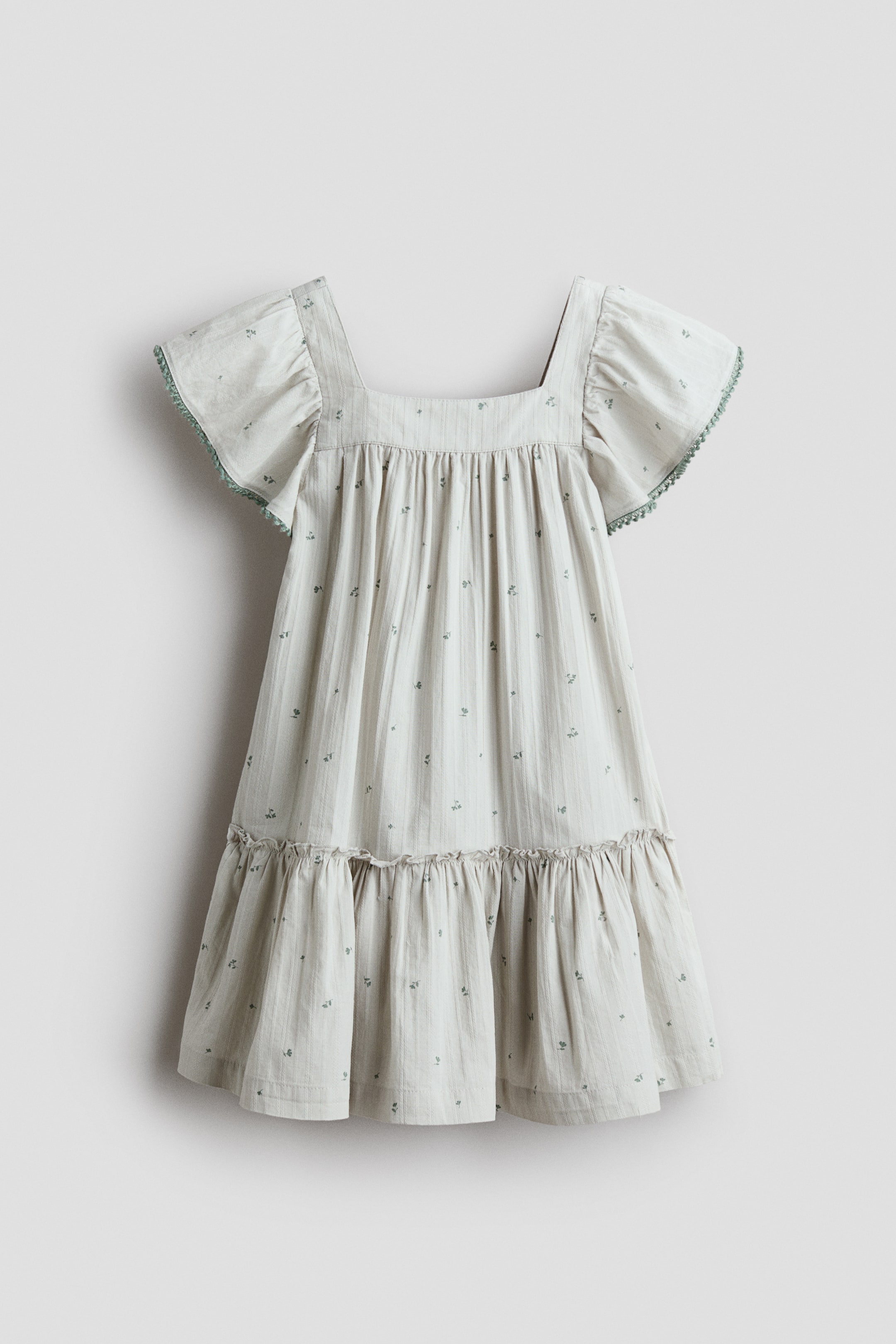 Visualizza immagine più grande: La PARTE ANTERIORE di un abito svasato presenta uno scollo quadrato e maniche a farfalla con bordo verde. È caratterizzato da un tessuto di cotone leggero con sottili righe verticali e piccoli motivi floreali verdi, insieme a una cucitura arricciata intorno alla parte superiore e un livello con volant rifinito sullo gonna.