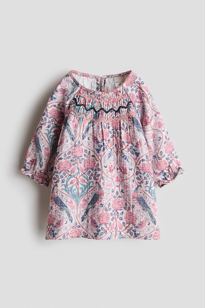 An A-line cotton muslin dress is shown from the front, featuring an all-over floral and bird print in pink, blue and green. The dress features a round neckline, a smocked chest panel with embroidered detailing, and long raglan sleeves with elasticated, frilled cuffs. - 1