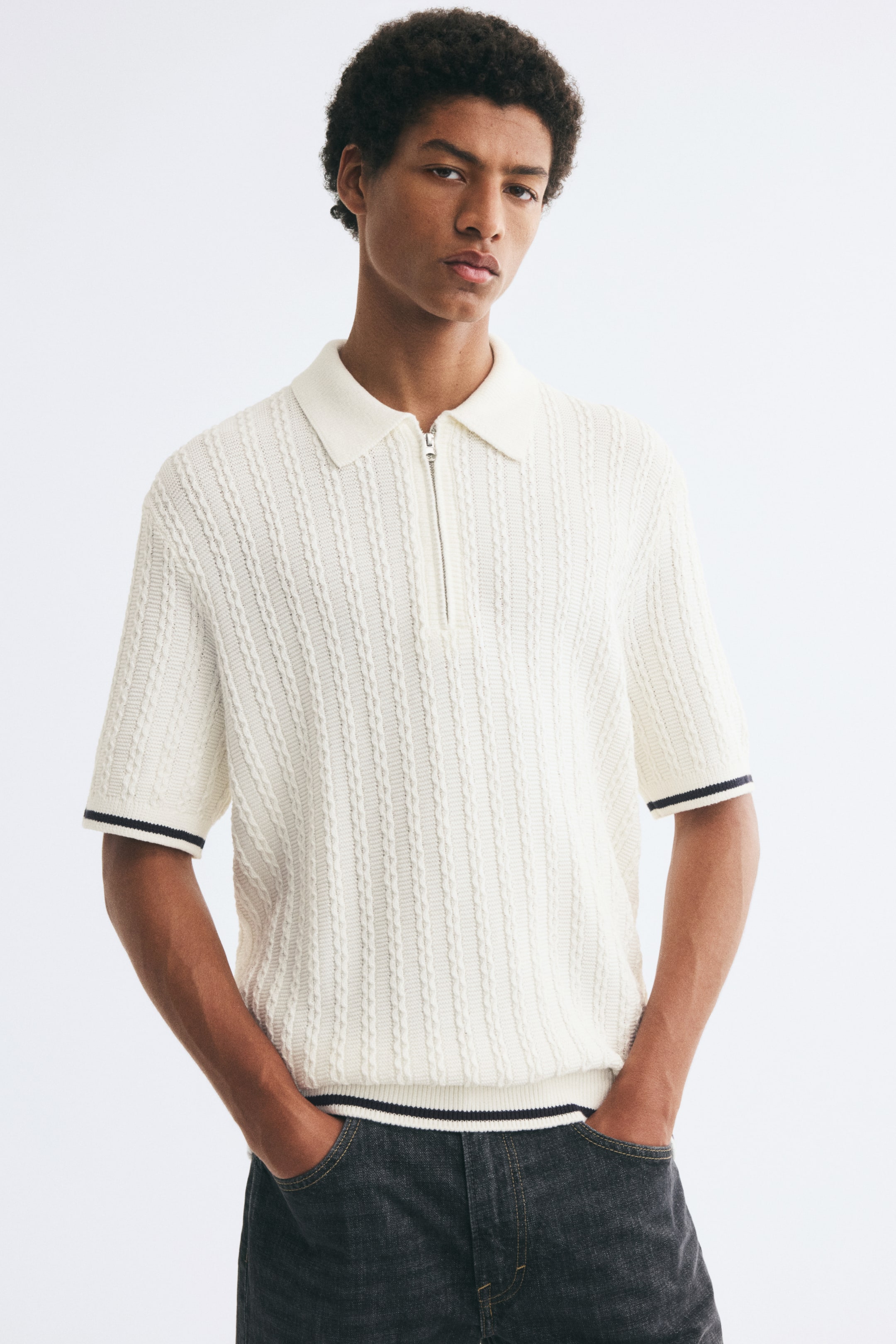 Xem ảnh lớn hơn: Mẫu áo polo tay ngắn bằng chất liệu dệt kim cable knit màu kem, có bề mặt kết cấu, đang được người mẫu mặc, nổi bật với cổ rib dệt gân, khóa kéo nửa cổ và các sọc tương phản màu tối ở bo tay và lai áo. Áo polo phom relaxed-fit được phối cùng quần jeans màu xám đậm, người mẫu đặt tay trong túi quần.