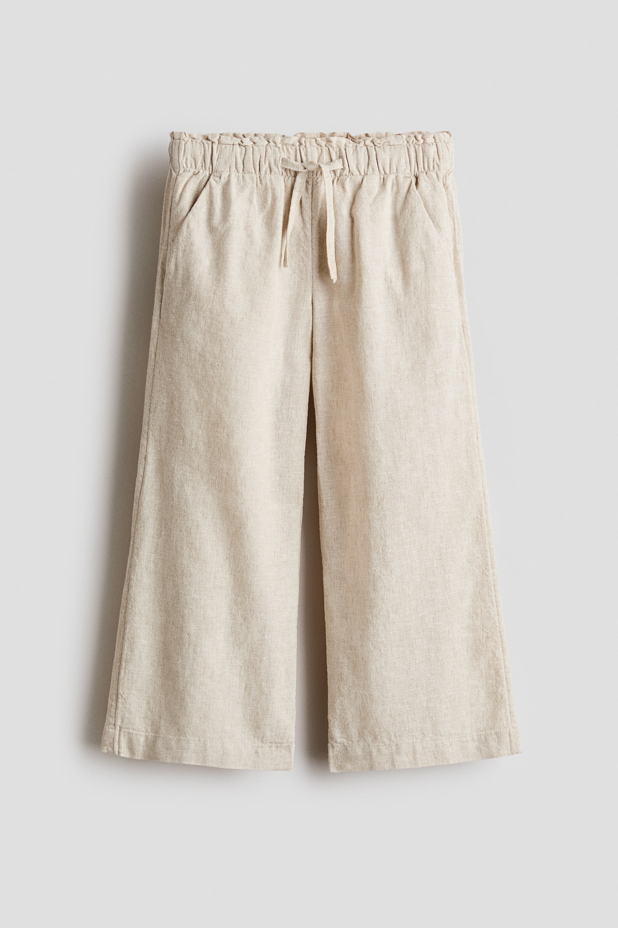View larger image: A pair of light beige wide-leg pants features an elasticized, ruched waistband with a drawstring and side-seam pockets. The cropped pants display a linen-like texture on the front.