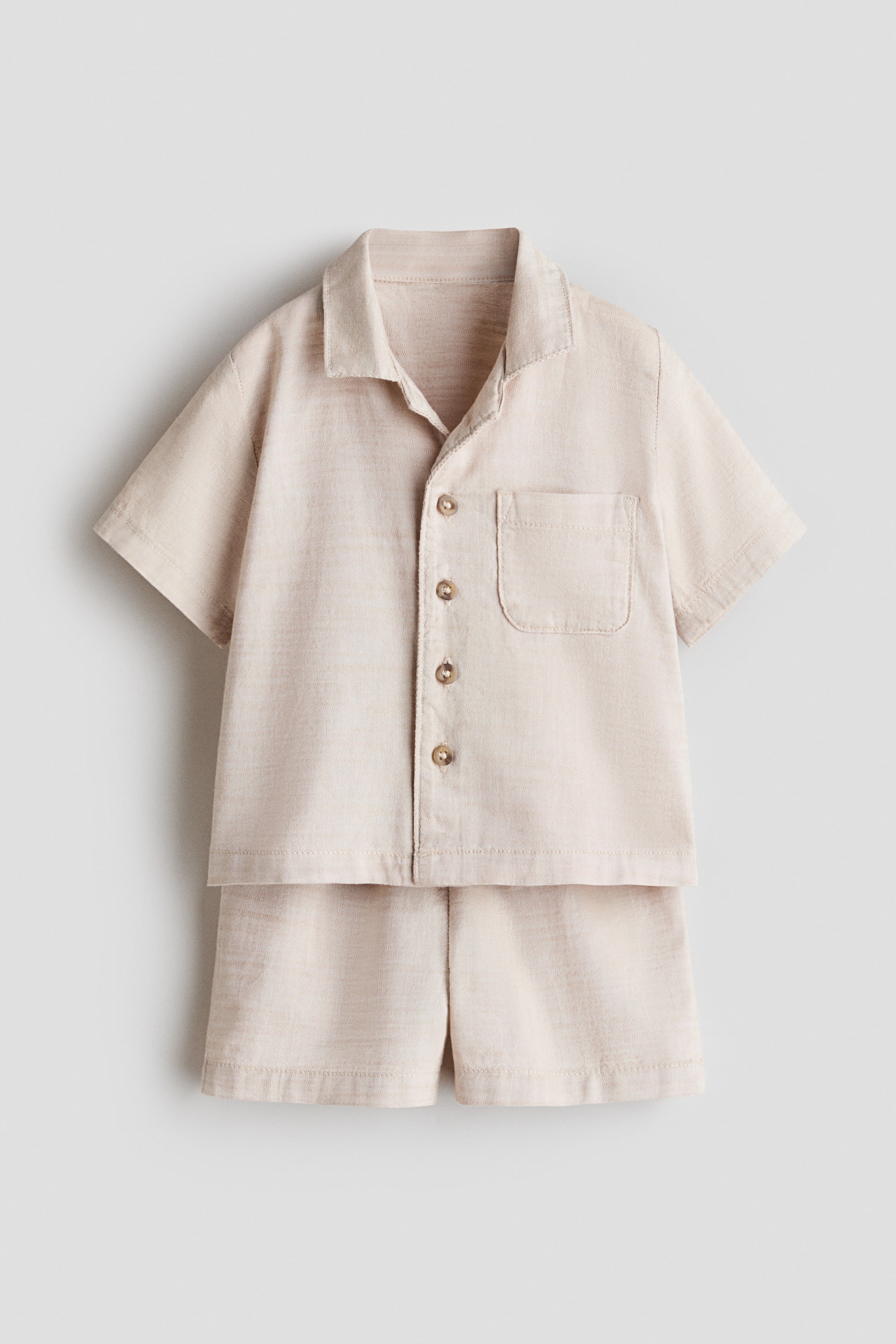 View larger image: A set of beige woven fabric shirt and shorts is appearing front-facing. The straight-cut shirt is featuring a resort collar, buttons down the front, short sleeves, and an open chest pocket, while the matching shorts are displaying an elasticated waist.