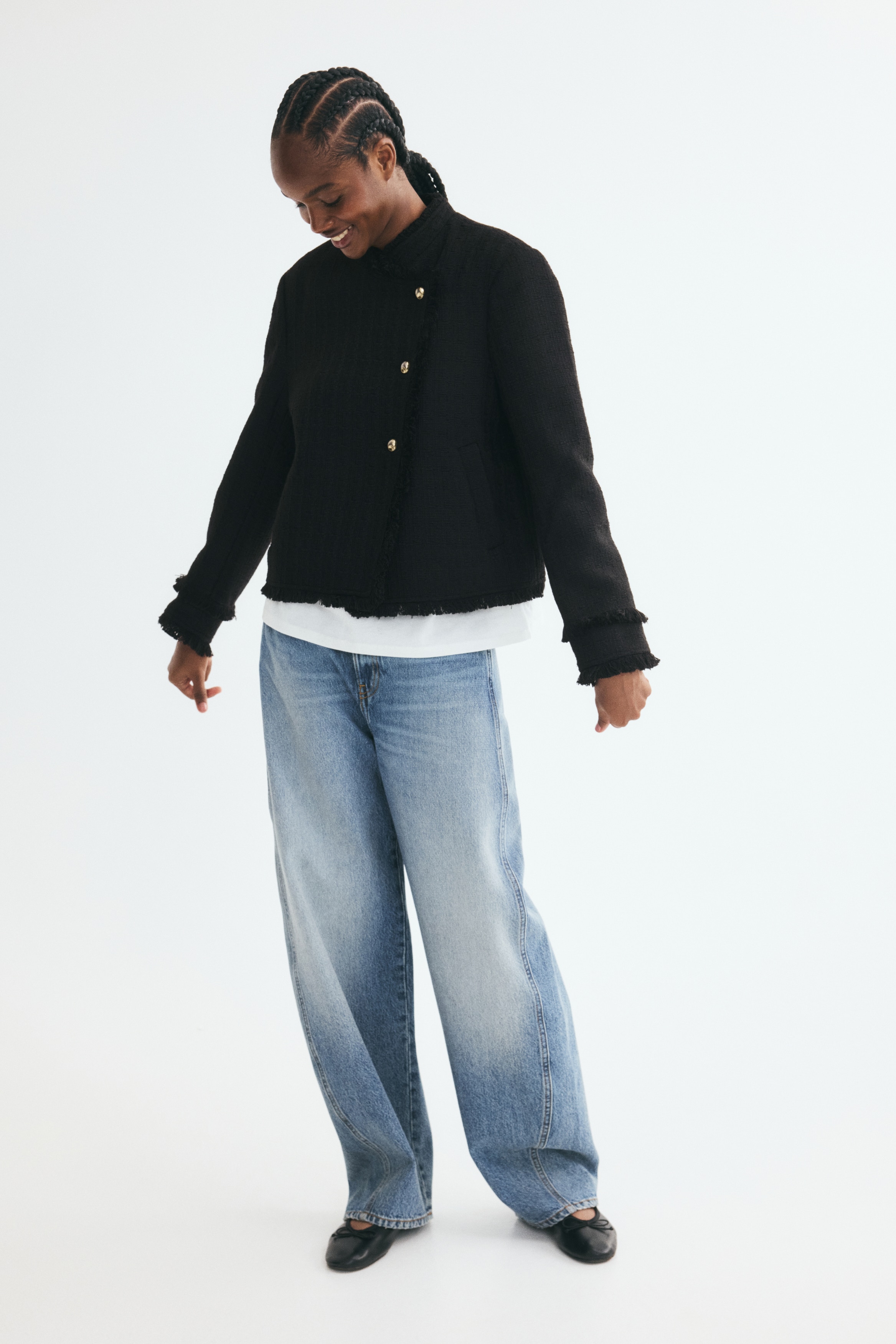 A black textured tweed jacket is shown from the front, featuring an asymmetrical closure secured by three gold-tone buttons and a standing collar. The jacket is detailed with frayed fringe trim along the collar, front opening, cuffs, and hem, and includes subtle side welt pockets.