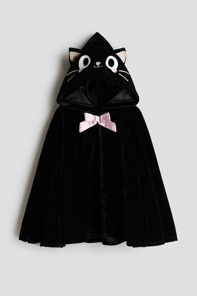 Une cape en velours noir présente une capuche en forme de tête de chat avec des yeux blancs, un nez noir, des moustaches brodées en or et des oreilles intérieures pointues dorées. Un nœud en satin rose orne l'encolure avant. - 1