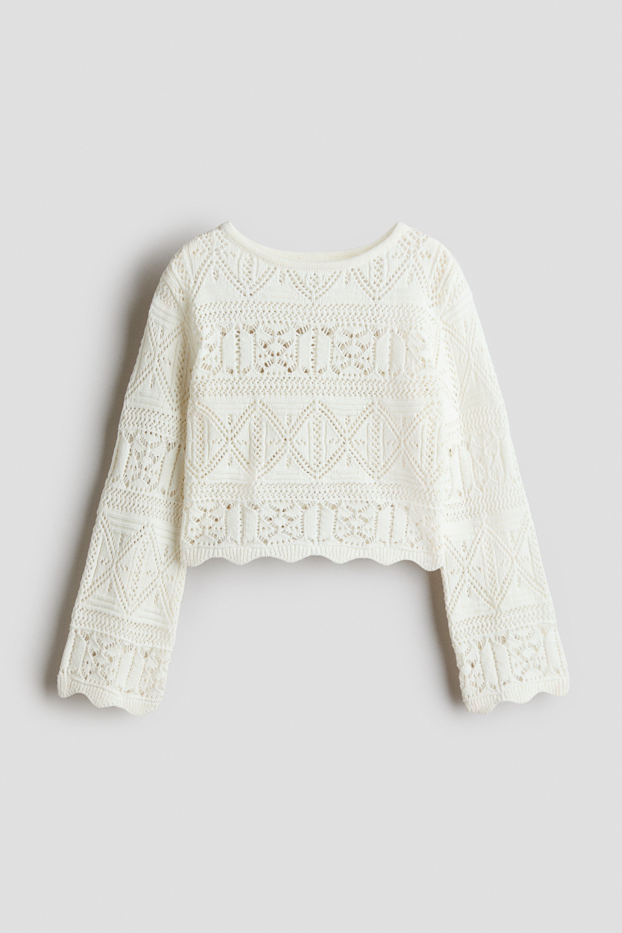 View larger image: A cream open-knit cropped sweater is showcased from the FRONT, highlighting detailed geometric patterns of diamonds and vertical bars across its body and long sleeves, completed with a scalloped hem.