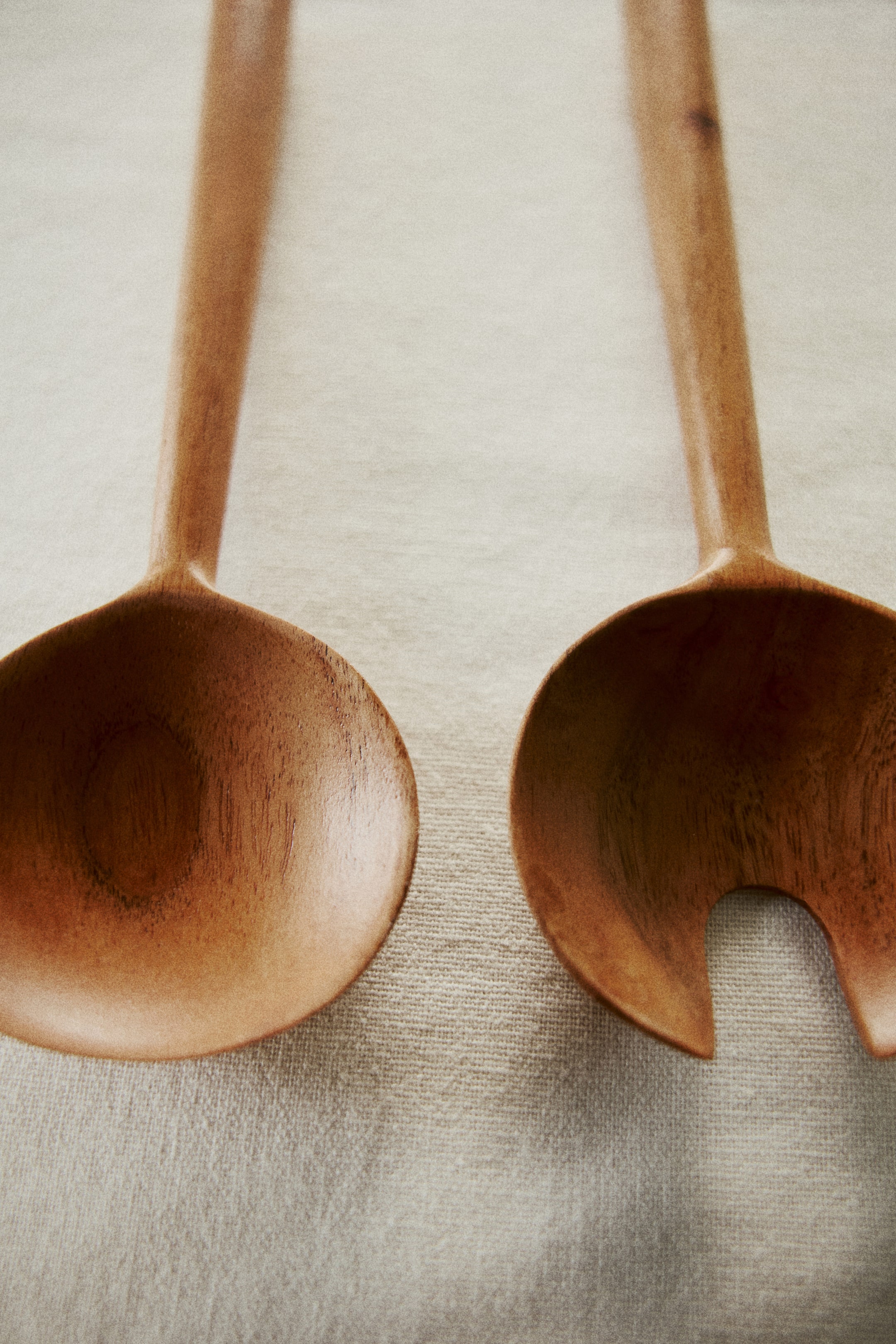 View larger image: A pair of wooden salad servers are lying side by side, angled towards the top. One server is a solid, deep spoon, and the other is a coordinating server featuring a large U-shaped cut-out for serving. Both display visible wood grain.