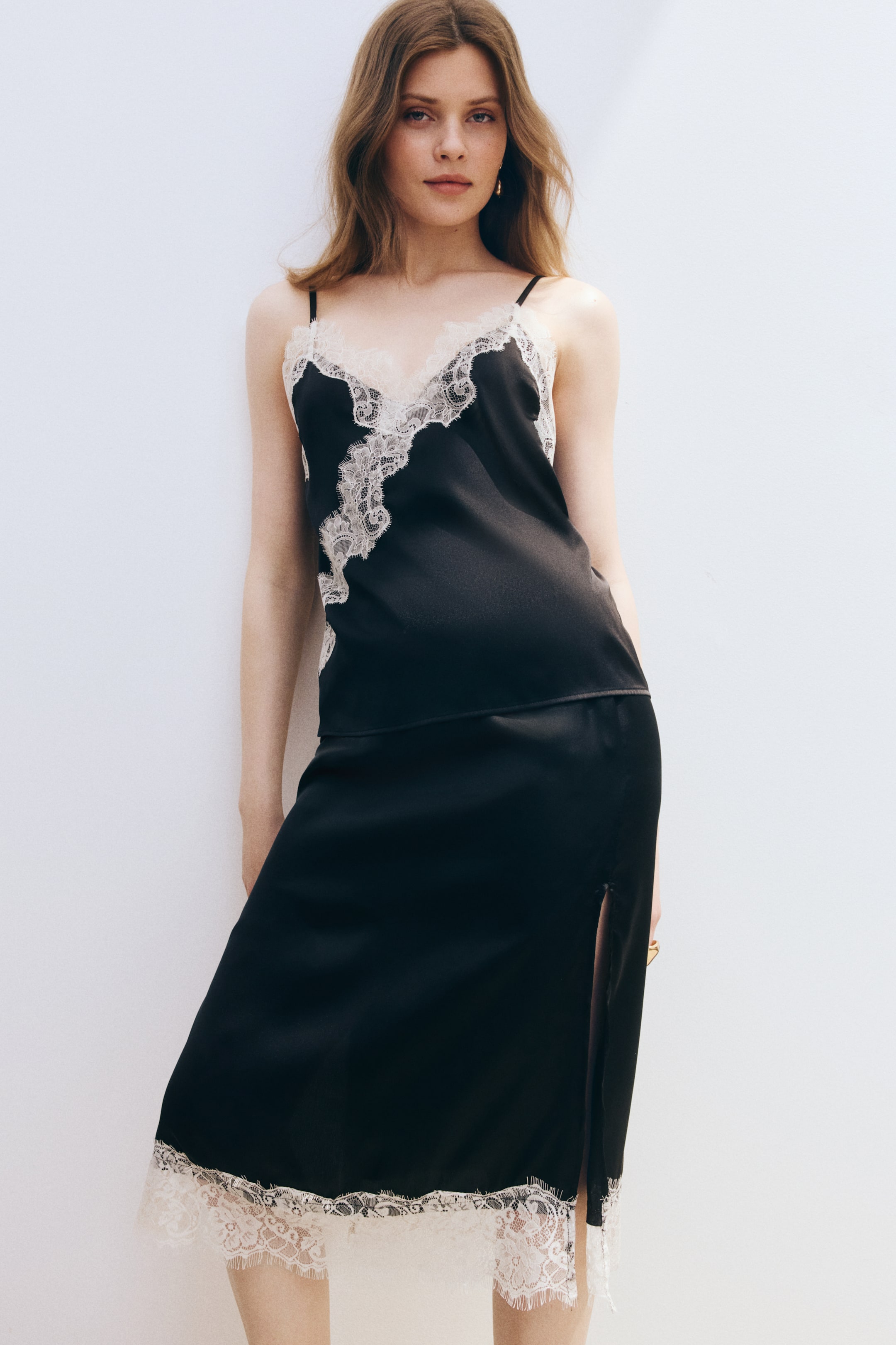 View larger image: A black slip dress featuring a V-neckline and thin spaghetti straps, showcasing intricate white lace trim along the neckline and a wide band of matching lace at the midi-length hem with a side slit.