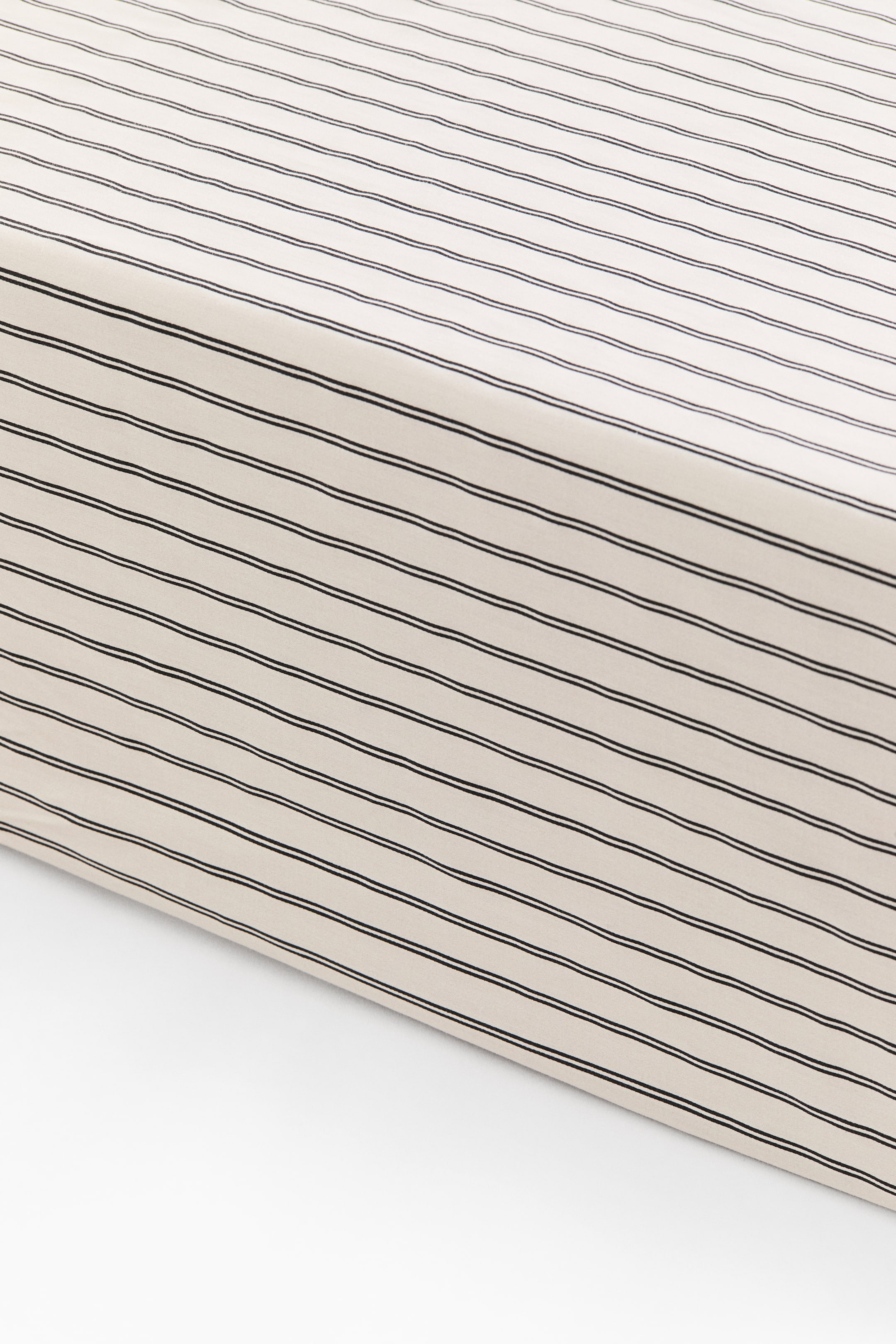 Agrandir l'image: Un drap-housse à motif recouvre le coin d’un matelas, démontrant un ajustement lisse et bien tendu. Le drap en satin de coton présente une surface beige clair, agrémentée de fines rayures parallèles foncées courant le long du dessus et des côtés.