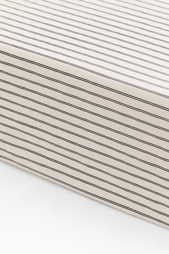 A patterned fitted sheet is covering the corner of a mattress, demonstrating a smooth and snug fit. The cotton satin sheet features a light beige surface detailed with thin, dark parallel stripes running along the top and sides. - 1