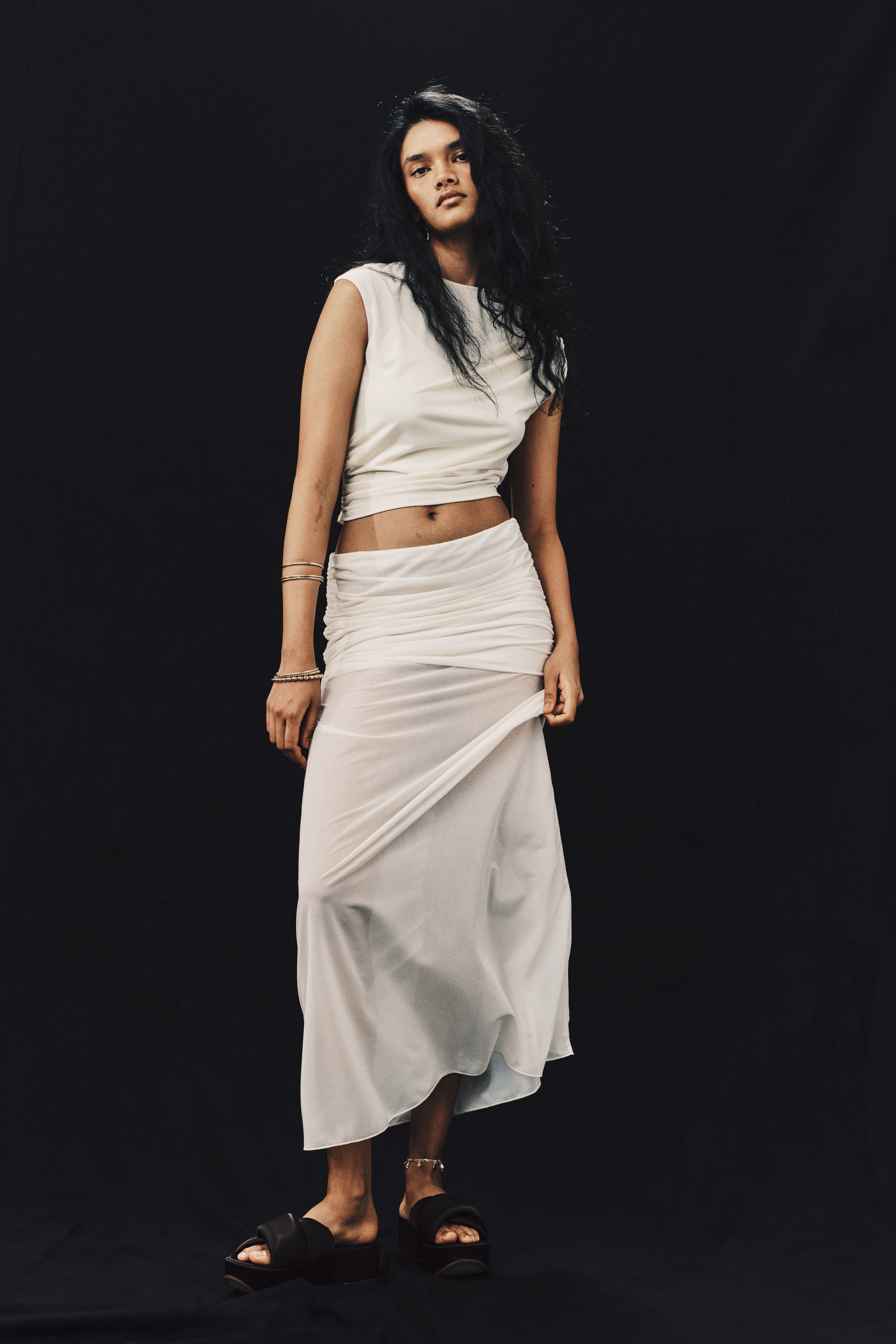 View larger image: A white jersey maxi skirt featuring a wide, gathered layer at the hips that creates a draped effect is worn by the model. The model is holding the side of the semi-sheer, flared hem of the skirt, styled with a matching sleeveless top.