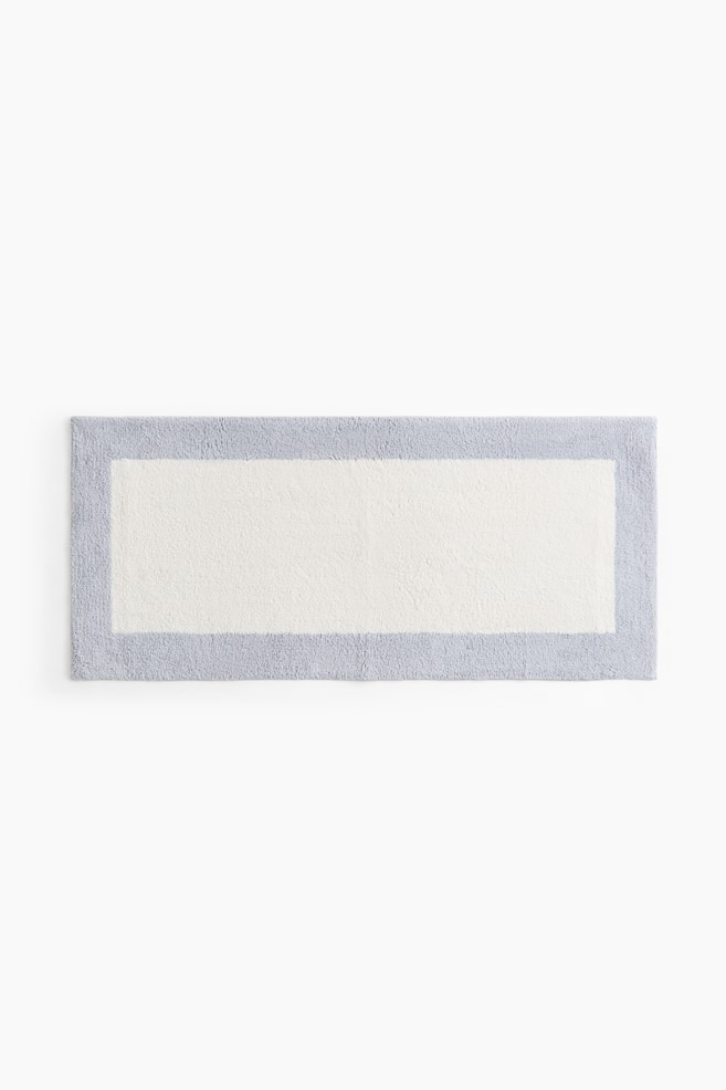 A rectangular tufted cotton bath mat is presented from the front, featuring an off-white central panel framed by a light blue border. - 1