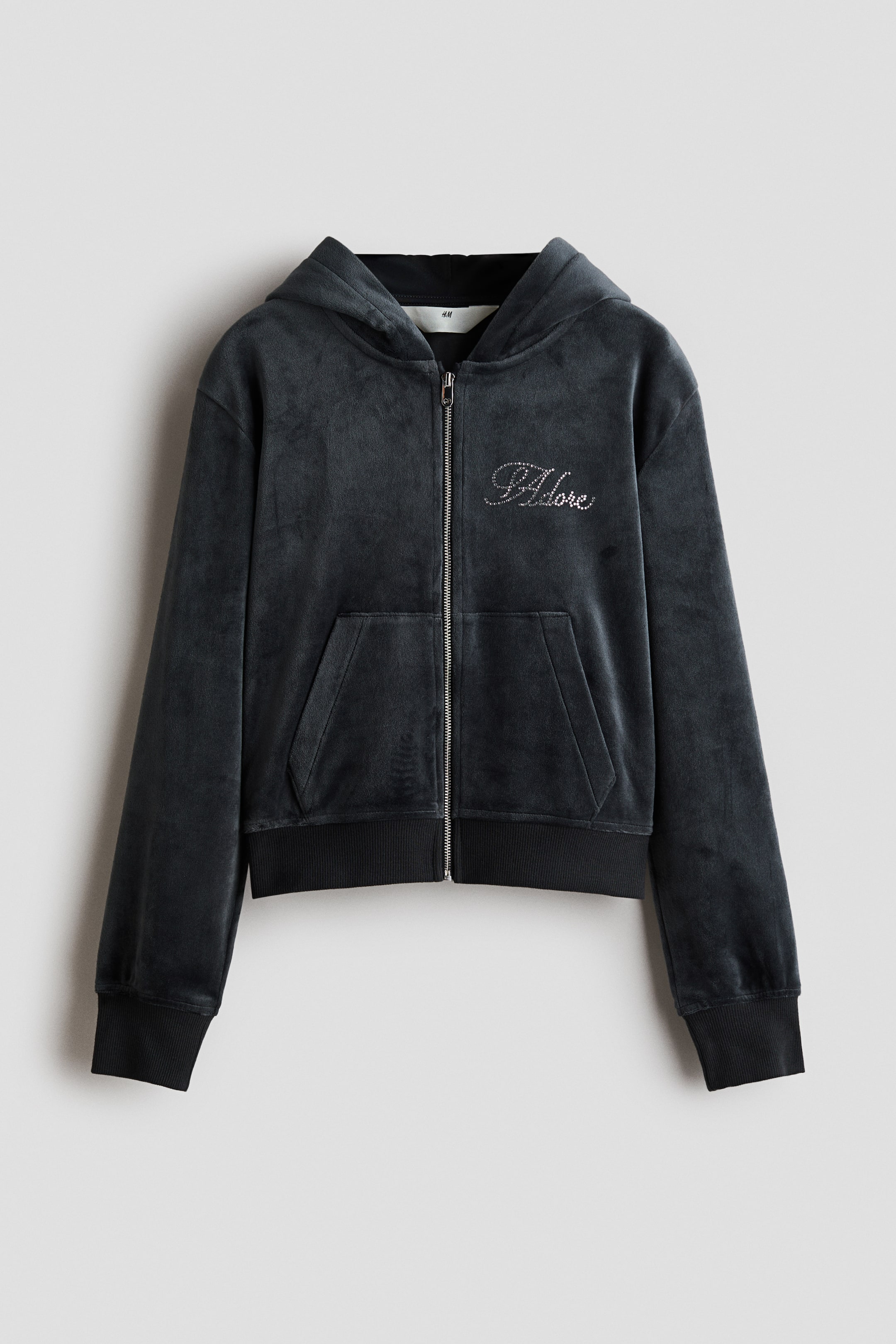 View larger image: A dark grey velour zip-up hoodie is shown from the front, featuring an attached hood, a full-length front zipper, and a split kangaroo pocket. A rhinestone-embellished cursive script details the left chest, while ribbed knit bands finish the cuffs and hem.