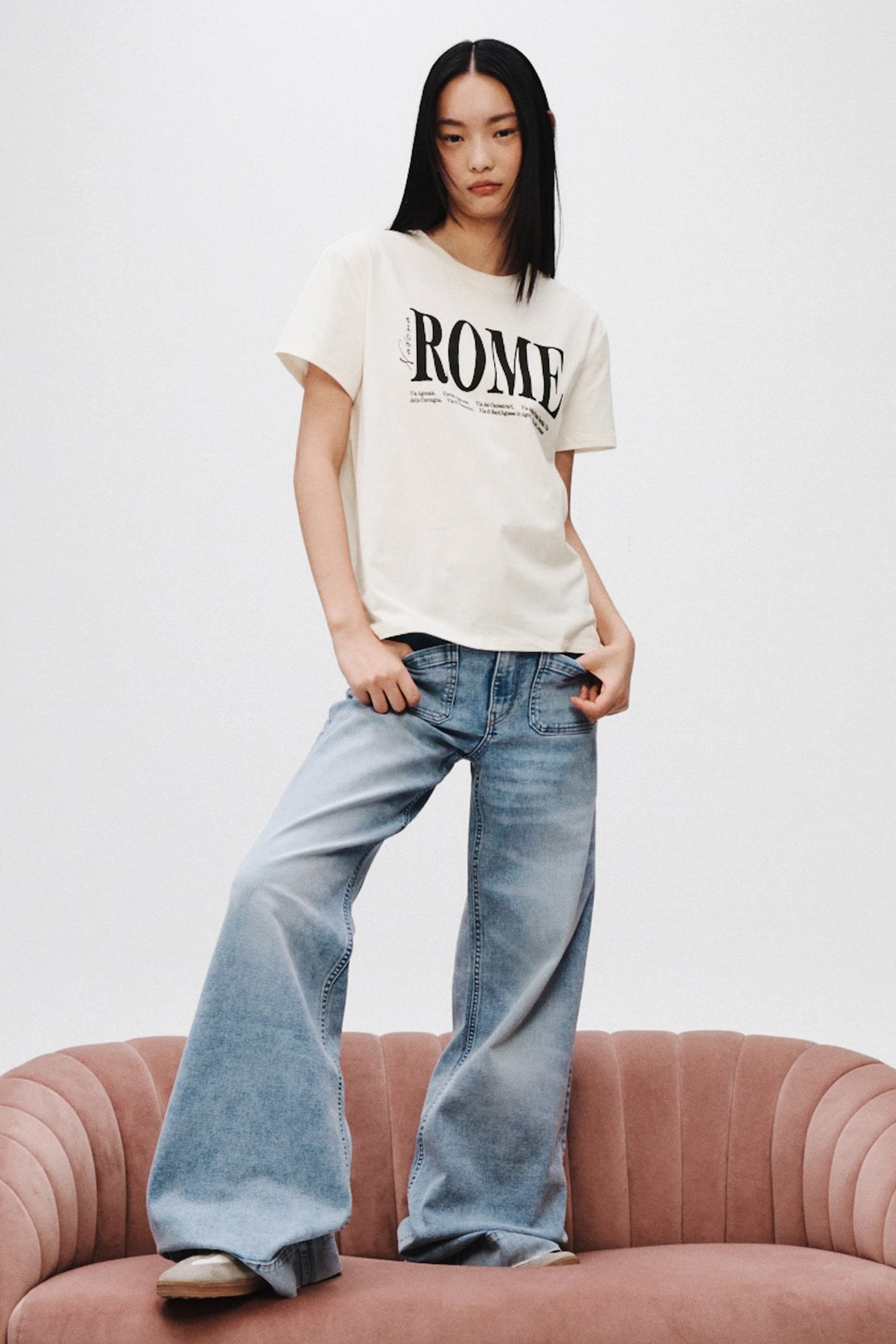 View larger image: A cream t-shirt displays 'ROME' in black lettering across the chest, its front tucked into light-wash denim jeans. The jeans feature a wide-leg, flared silhouette with a subtle ombré effect and visible front patch pockets.