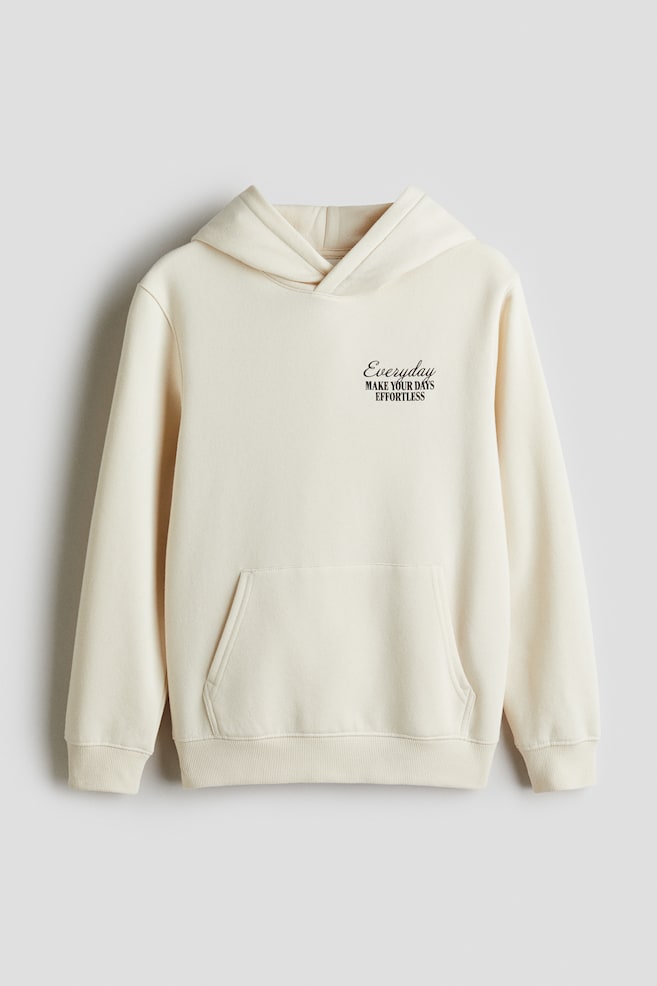 A cream-colored hoodie made from cotton sweatshirt fabric is shown from the FRONT, featuring a lined hood with a wrap front, dropped shoulders, and a kangaroo pocket. It is finished with ribbed cuffs and a ribbed hem, and is displaying black text that reads 'Everyday MAKE YOUR DAYS EFFORTLESS' on the left chest. - 1
