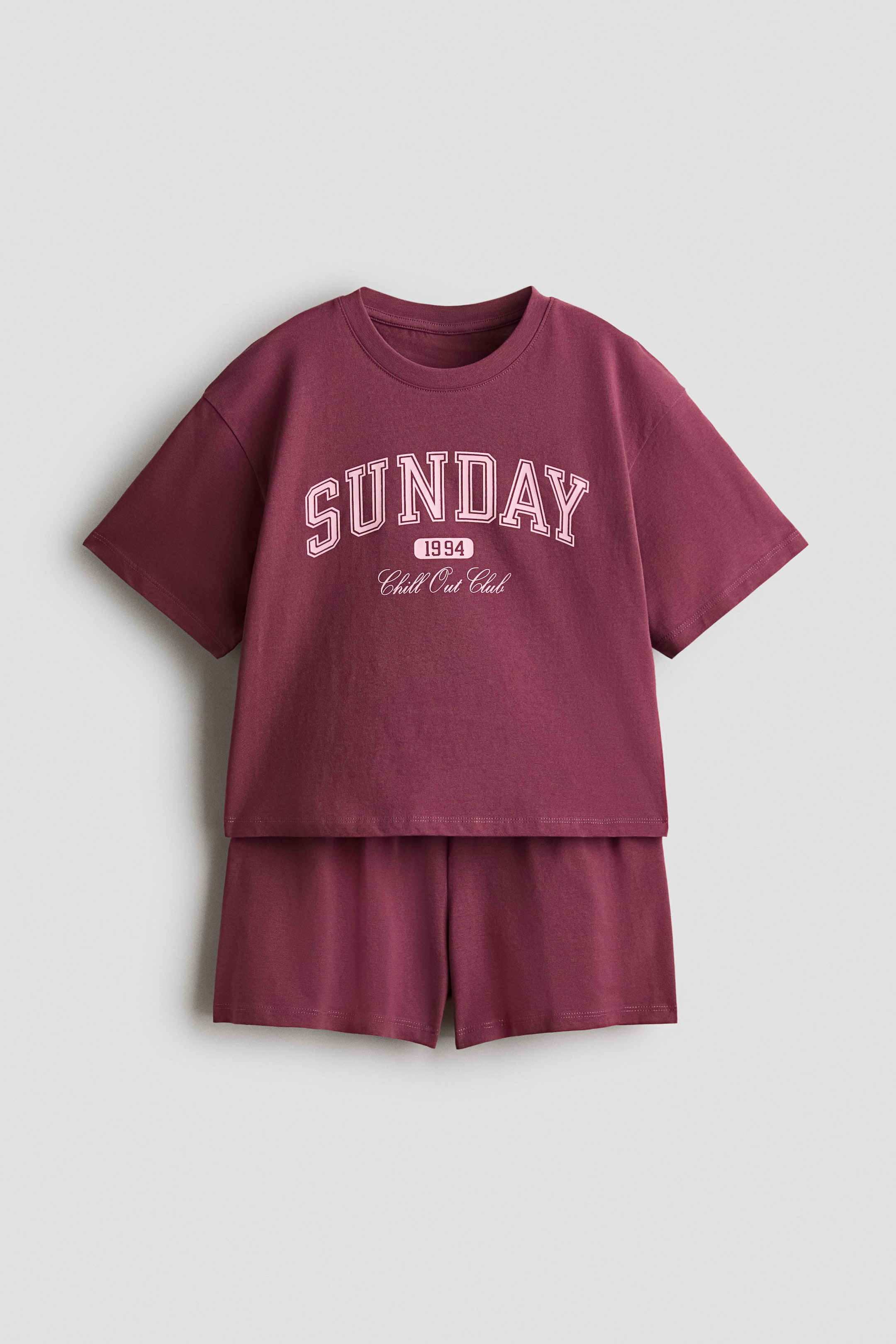 拡大画像を表示: 前面から見たマルーンの2ピースルームウェアセットは、クルーネックの半袖Tシャツとマッチングショーツを特徴としています。Tシャツには、アーチ状の淡いピンクのブロック文字で「SUNDAY」、その下に「1994」、そして胸には筆記体で「Chill Out Club」と表示されています。