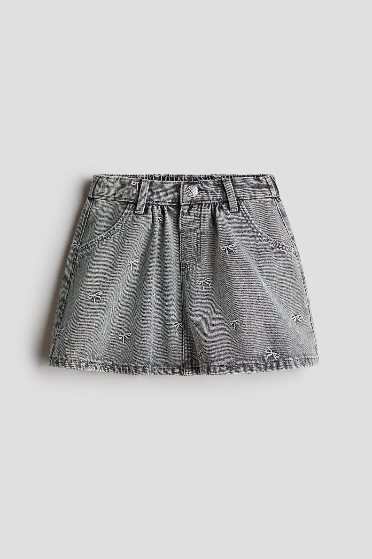 Een grijze denim rok van voren, met een all-over patroon van kleine, geborduurde witte strikjes. De rok heeft een elastische tailleband met riemlussen, een centrale metalen knoopsluiting en afgeronde steekzakken aan de voorkant.