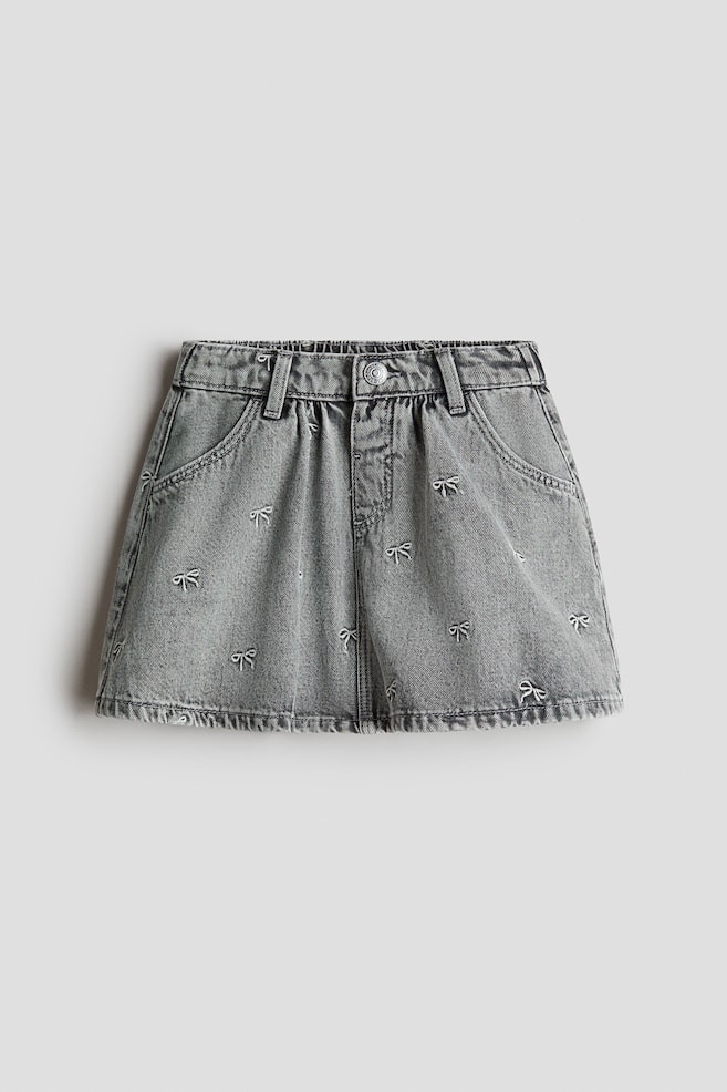 Ein vorne sichtbarer grauer Jeansrock mit einem Allover-Muster aus kleinen, aufgestickten weiÃen Schleifen. Der Rock verfÃ¼gt Ã¼ber einen elastischen Bund mit GÃ¼rtelschlaufen, einen mittigen Metallknopfverschluss und geschwungene Fronttaschen. - 1