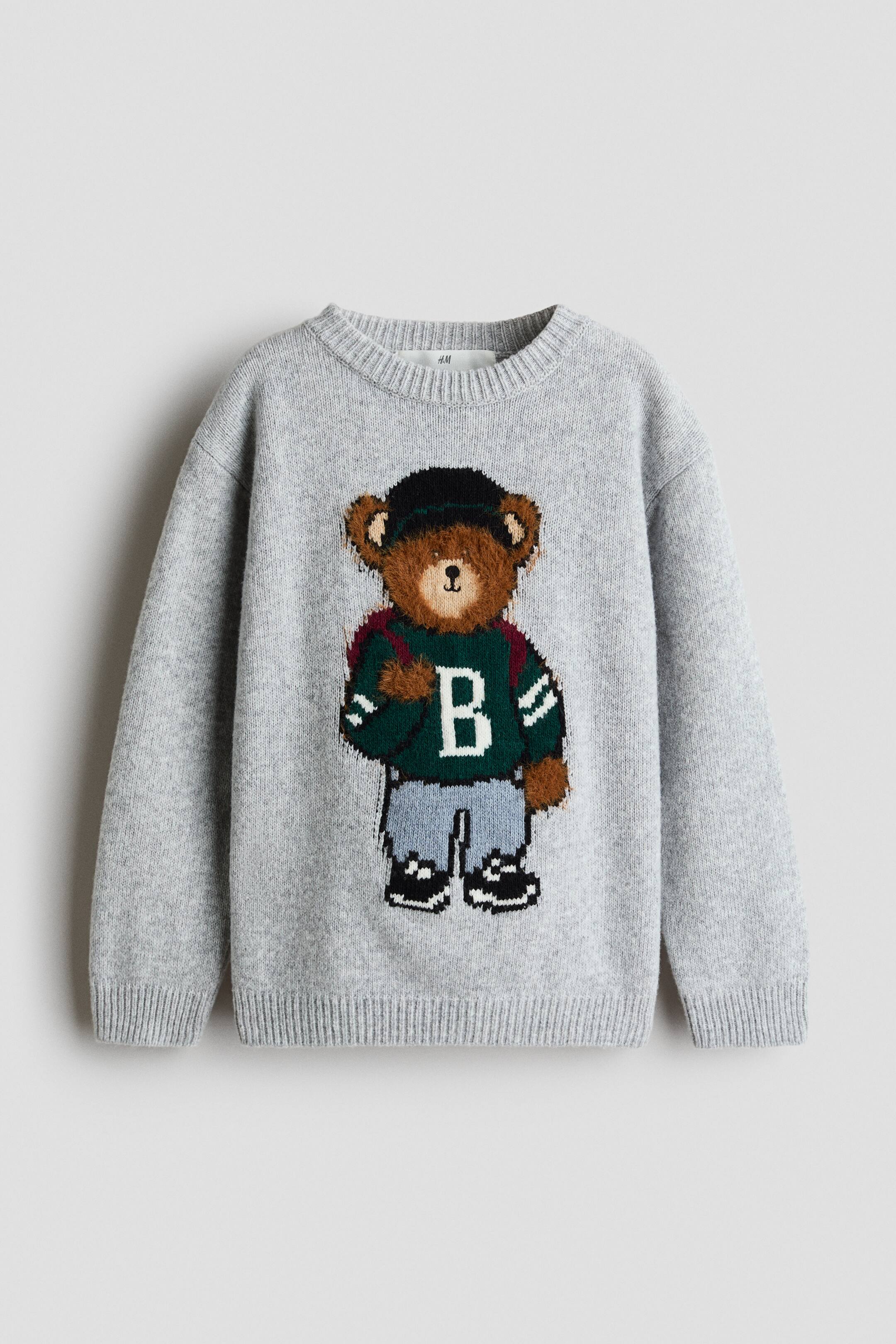 View larger image: A light grey knitted crew neck sweater showcases a brown bear character on its front, facing forward. The bear is adorned in a dark green varsity-style top with a white 'B', light blue trousers, black shoes, a dark cap, and a red backpack. The sweater is further accentuated with ribbed cuffs and hem.