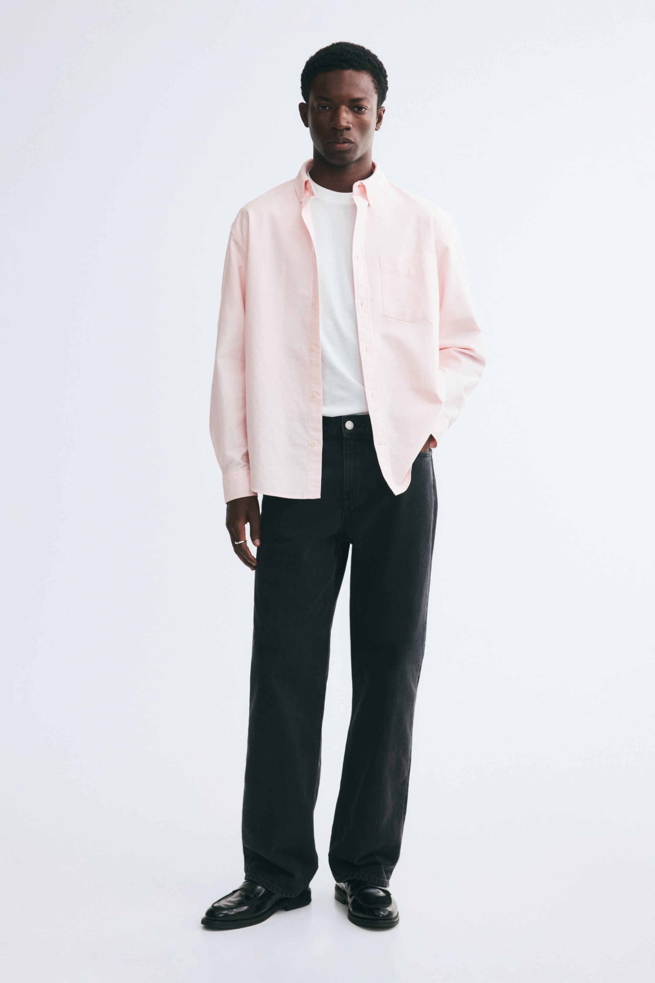 View larger image: A light pink Oxford cotton shirt is worn unbuttoned by the model over a white T-shirt and black jeans. The shirt features a button-down collar, a single open chest pocket, and long sleeves.