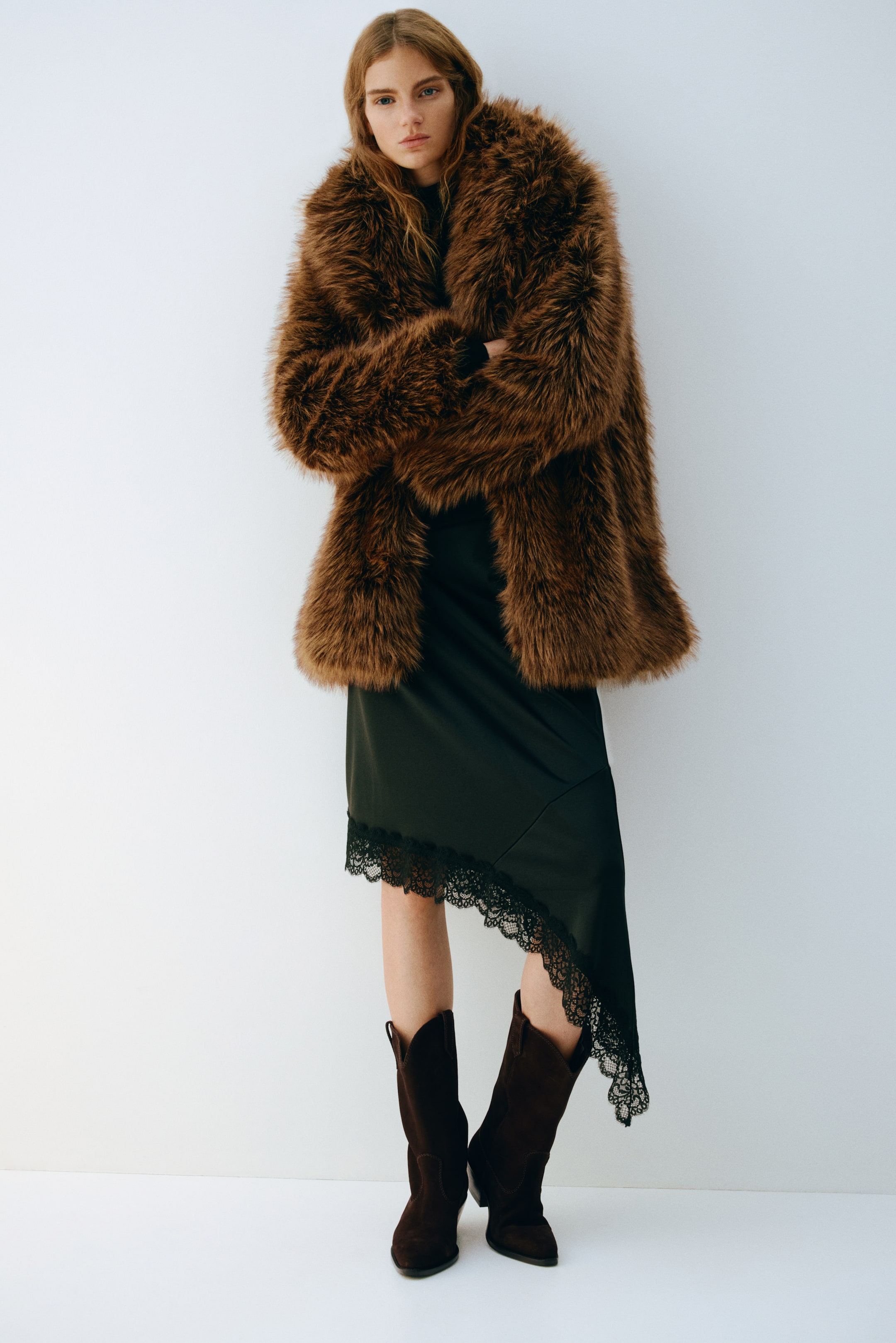 View larger image: A brown faux fur coat is featuring a shaggy, voluminous texture and an oversized silhouette, being worn with arms crossed and layering over a dark slip dress.