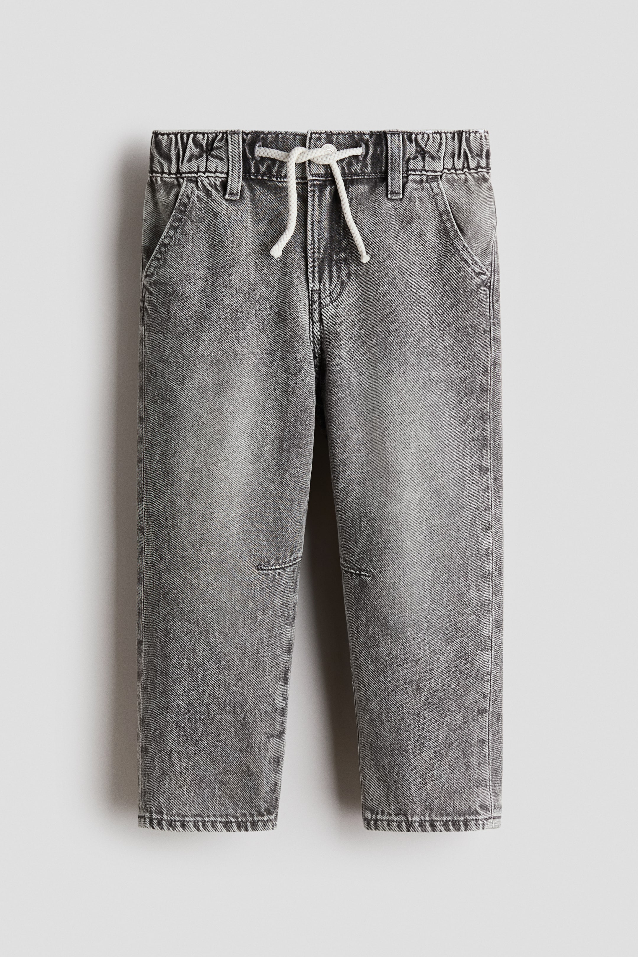 View larger image: Grey denim joggers are displayed from the front, featuring a relaxed fit through the hip and thigh with tapered legs. They showcase an elasticated waistband with a white decorative drawstring tied at the front, a visible fly closure with a button, side pockets, and darts on the knees.