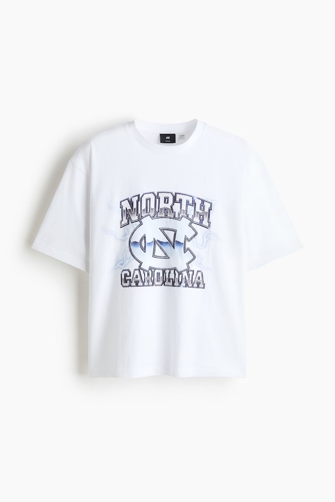 Ein weiÃes T-Shirt mit Rundhalsausschnitt ist von vorne zu sehen und zeigt eine lockere Passform sowie weite kurze Ãrmel. In der Mitte befindet sich ein Grafikdruck mit dem bogenfÃ¶rmigen Wort âNORTHâ Ã¼ber einem ineinander verschlungenen âNCâ-Logo und âCAROLINAâ darunter in einem strukturierten Blau-Grau-Muster. - 1