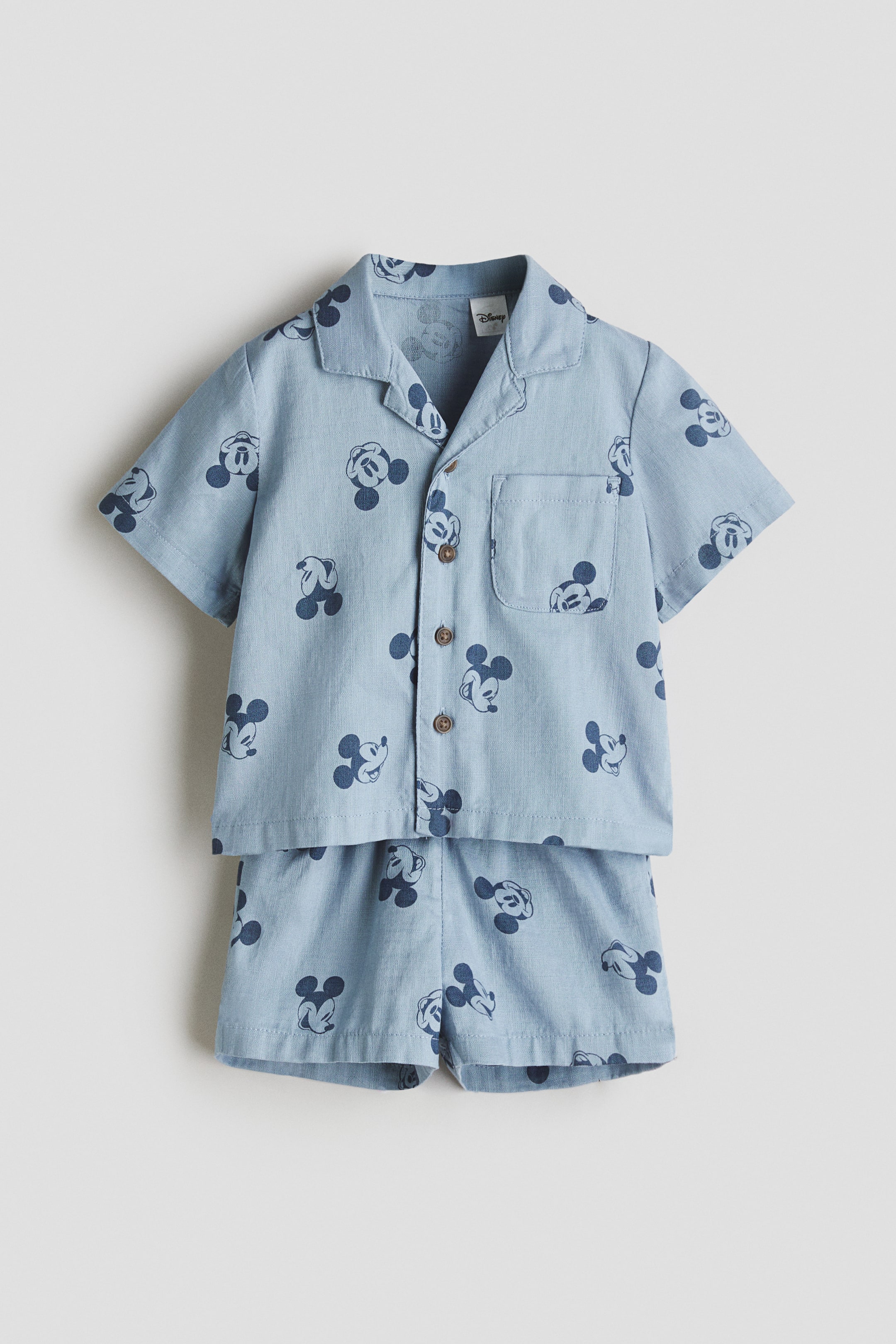 Voir une image plus grande: Un ensemble chemise et short en tissage de coton bleu clair est présenté de face, arborant un imprimé intégral de Mickey Mouse en bleu foncé. La chemise est conçue avec un col de style resort, des boutons sur le devant, une poche poitrine ouverte et des manches courtes avec revers cousus, tandis que le short affiche également des revers cousus aux ourlets.