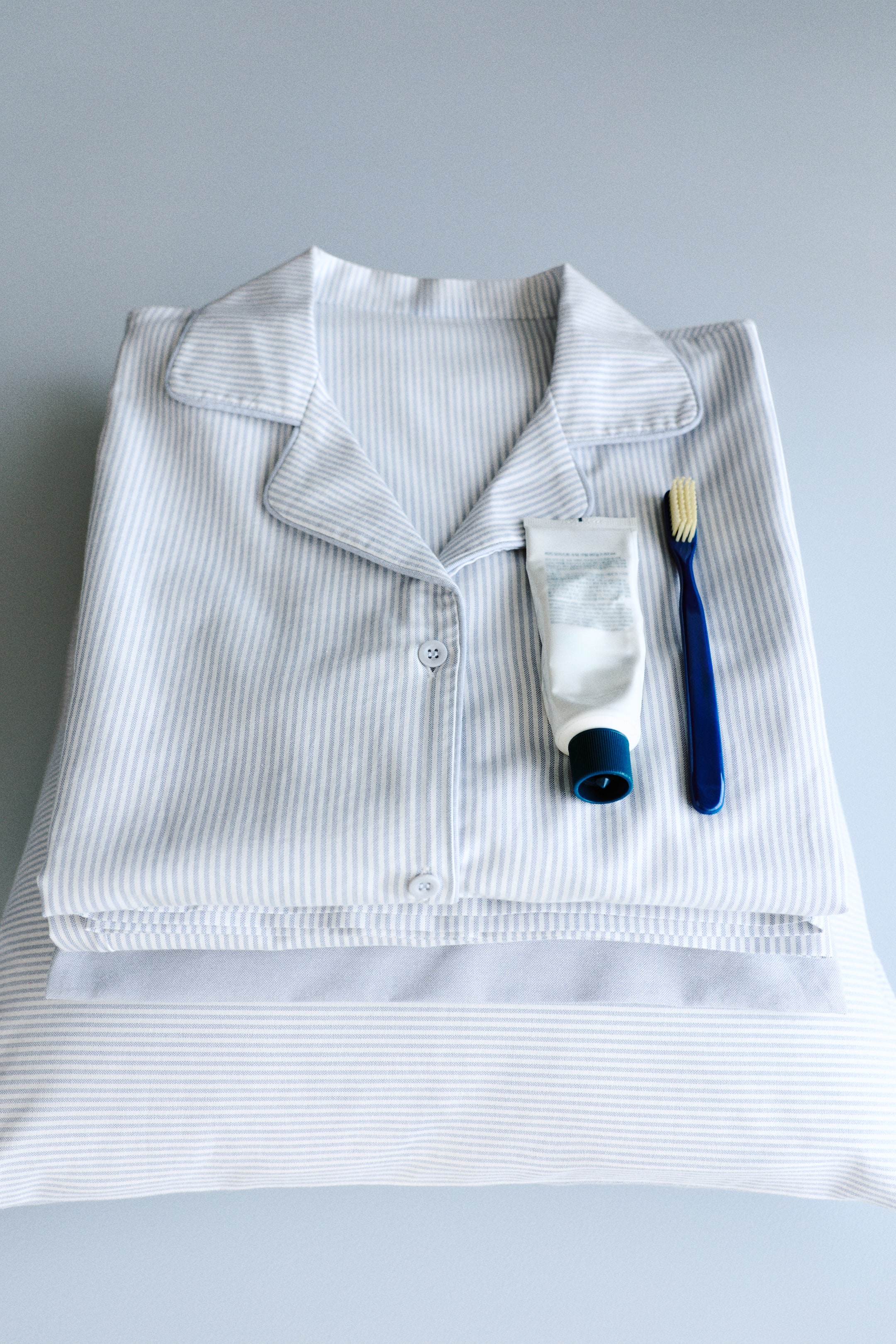 View larger image: A pyjama set is laid folded flat, showing its front, consisting of a shirt and bottoms in a light blue and white striped pattern. The shirt features a rounded resort collar, buttons down the front, and piping along the collar and button band.