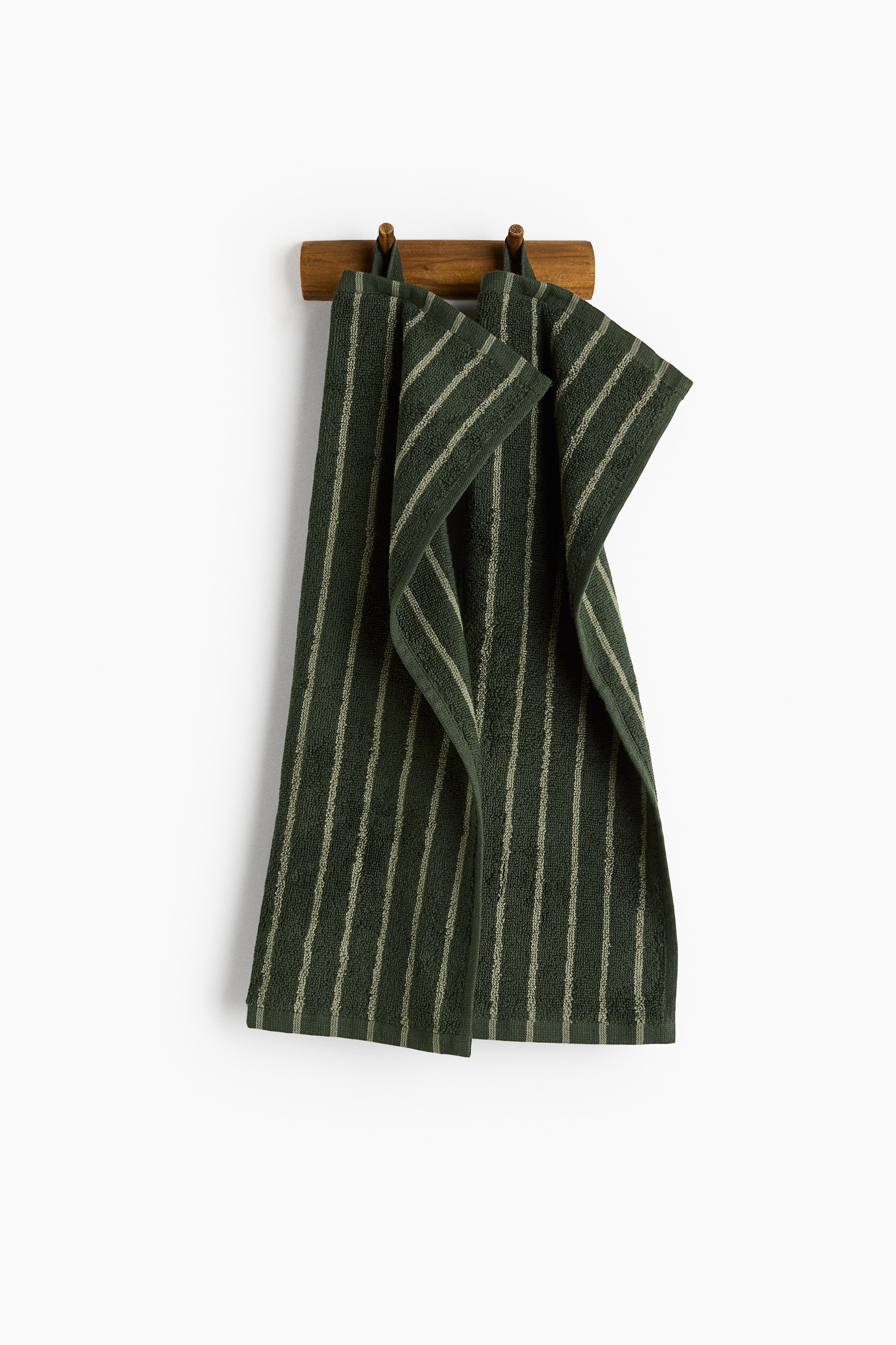 View larger image: Two dark green cotton terry guest towels featuring thin, light green vertical stripes are hanging from integrated loops on a wooden peg rack. The towels are draping downward, showcasing their soft, textured surface and woven bottom hems.