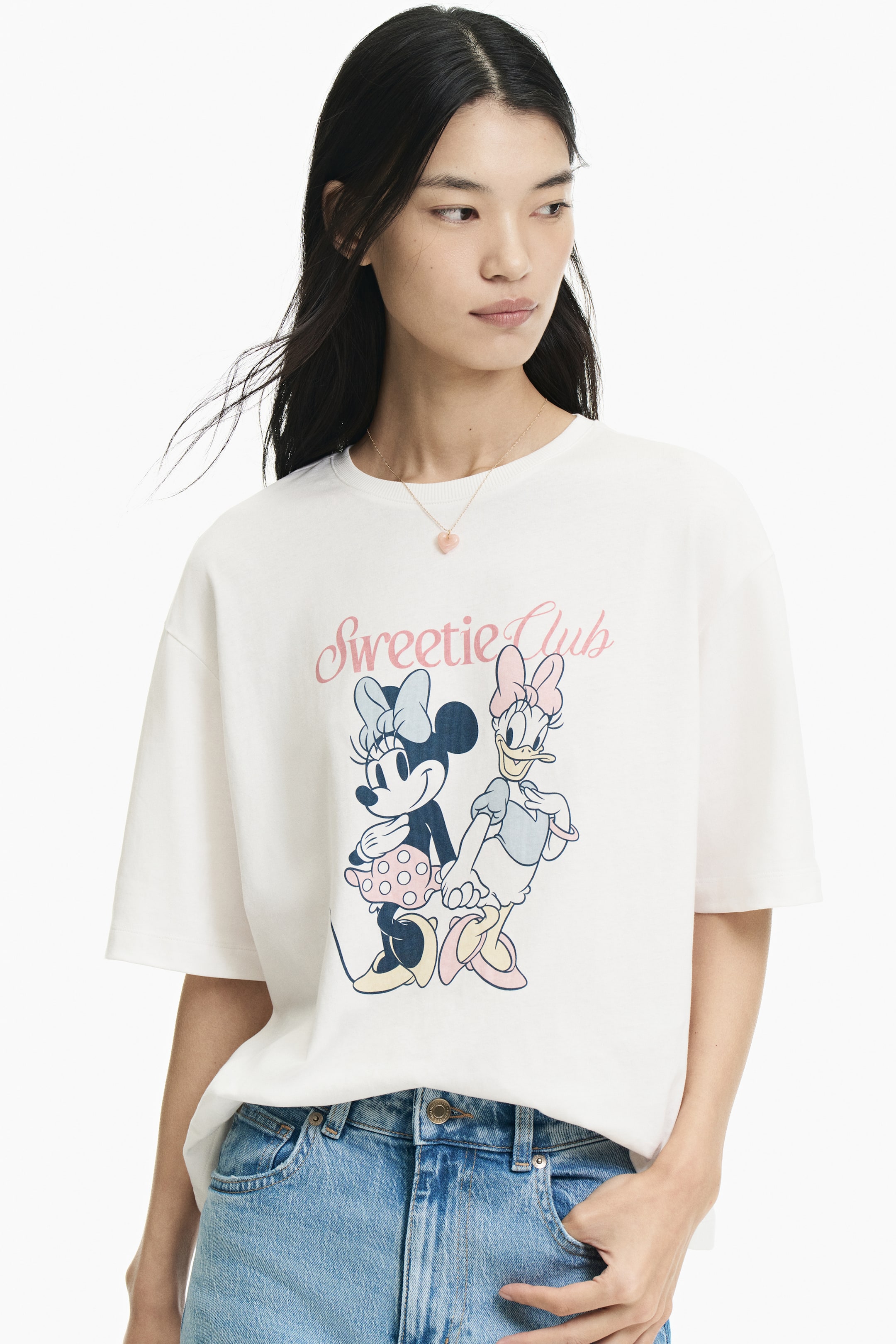 Xem ảnh lớn hơn: Người mẫu đang mặc một chiếc áo thun jersey cotton mềm màu trắng ngà. Sản phẩm nổi bật với cổ tròn bo gân, vai trễ, lai áo cắt thẳng và họa tiết in phía trước với dòng chữ "Sweetie Club" cùng hình Minnie Mouse và Daisy Duck nắm tay nhau. Áo thun được phối cùng quần jeans xanh, và một sợi dây chuyền mặt trái tim màu hồng đang đeo trên cổ áo.