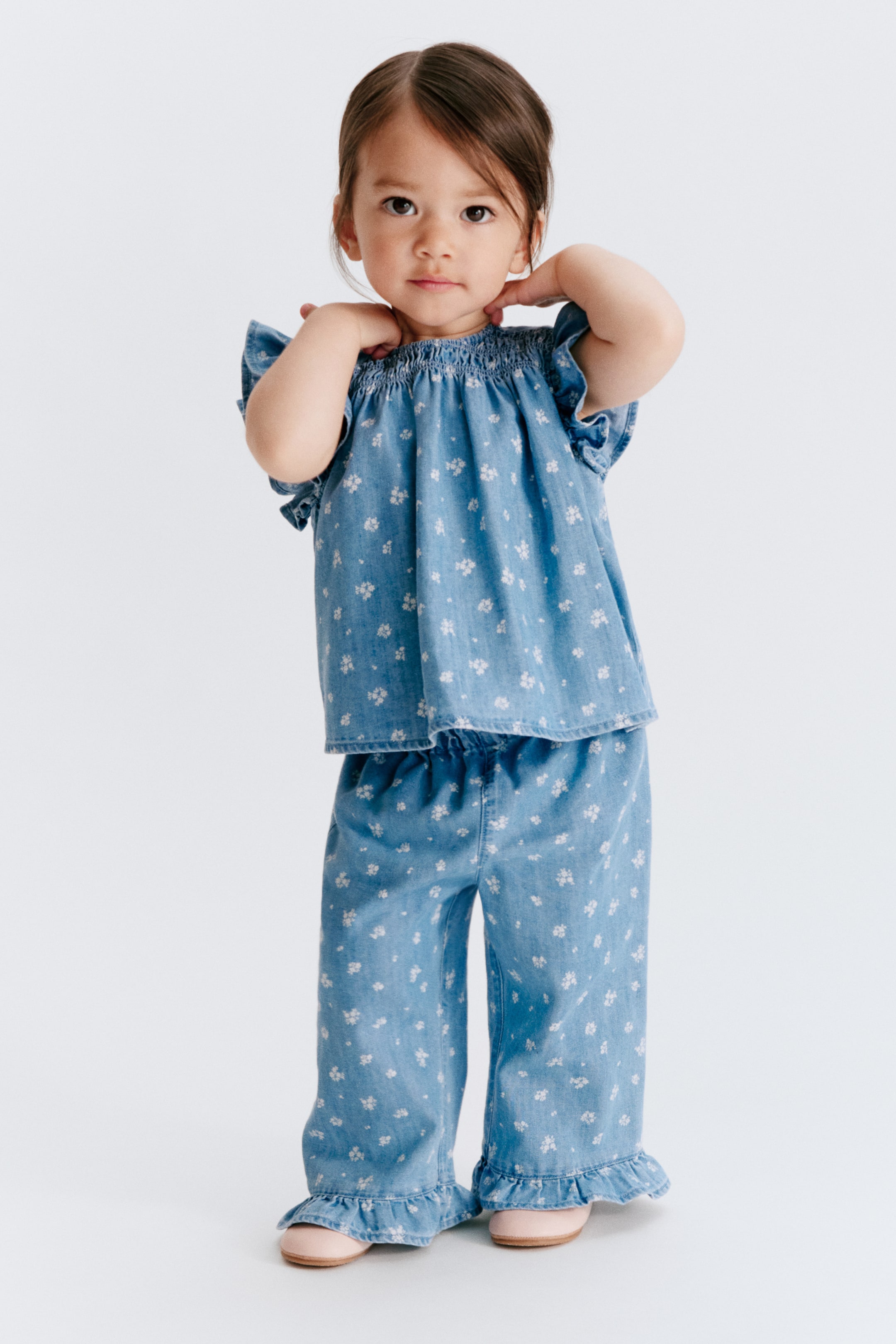 View larger image: A light blue two-piece children's outfit, crafted from a denim-look fabric, features a white floral pattern. The top is designed with a smocked neckline and ruffled cap sleeves, while the matching wide-leg pants showcase an elasticized waistband and ruffled ankle cuffs.