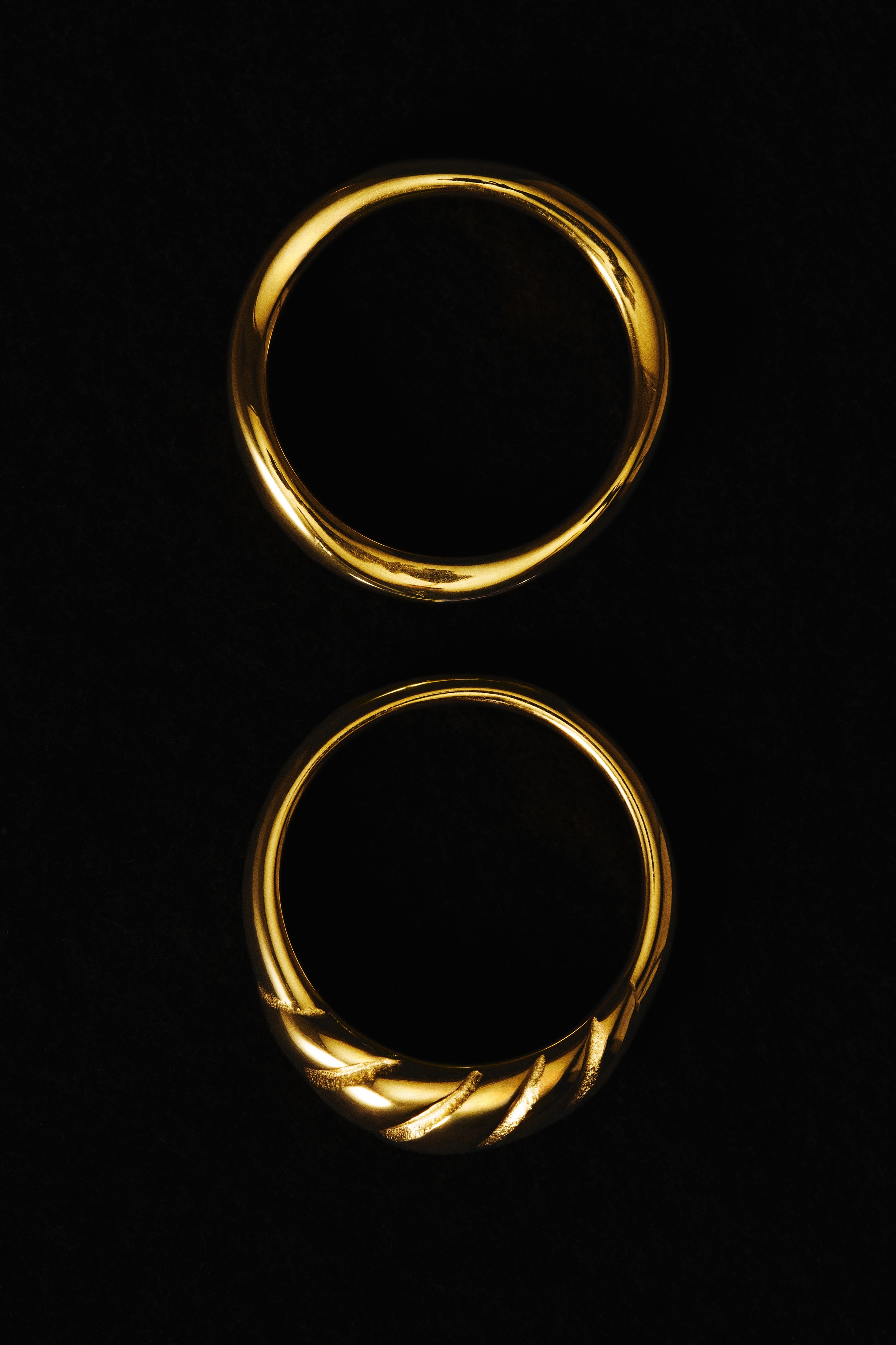 Two gold-toned rings are placed side by side, facing forward to showcase their polished finish. The ring on the left features a smooth, organic wavy band, while the ring on the right highlights a textured, ribbed croissant-style design on the front.