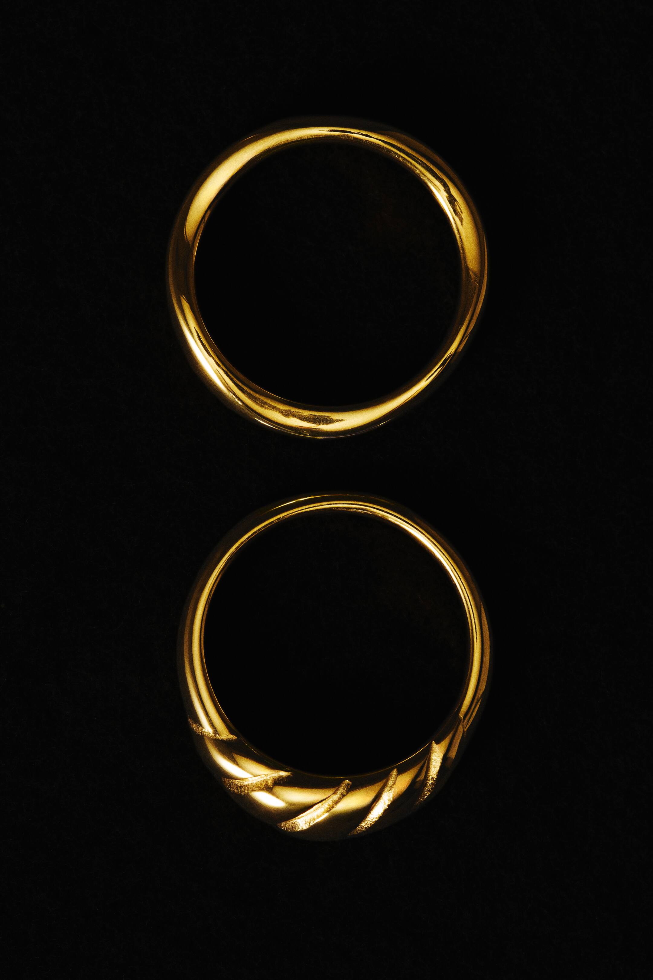 View larger image: Two gold-toned stainless steel rings are shown from the front, displaying different designs. The top ring features a polished, slim band with a subtle twist, while the bottom ring showcases a wider, domed band with textured diagonal grooves.