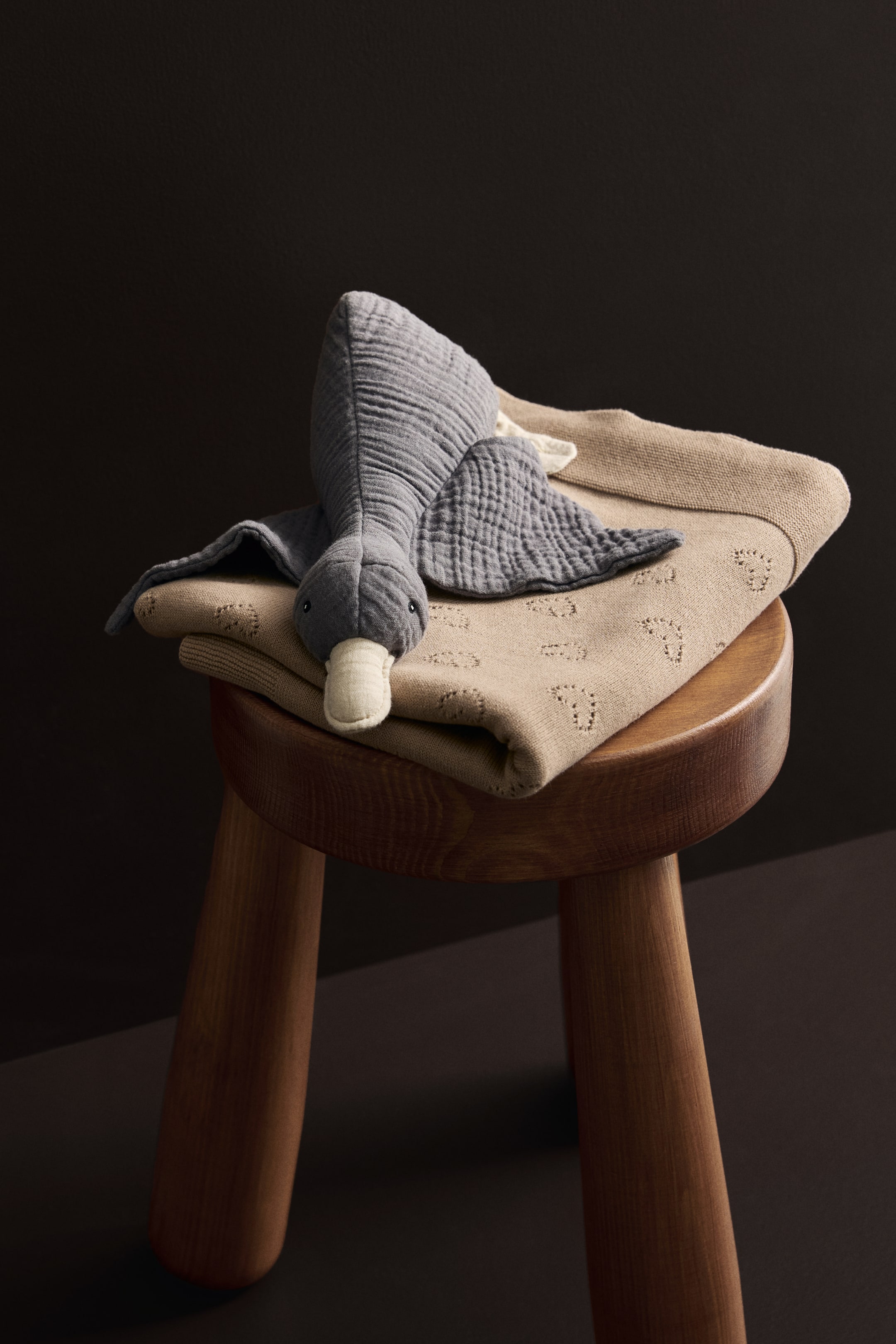 Zobacz większy obraz: Szara zabawka w kształcie gęsi leży na złożonym beżowym dzianinowym kocu. Zabawka gęś jest zwrócona w lewo, ma pomarszczone szare ciało, czarne oczy i jasny beżowy dziób, a jej skrzydła są rozpostarte. Beżowy koc prezentuje subtelny, powtarzający się wzór z nacięciami na całej swojej powierzchni.