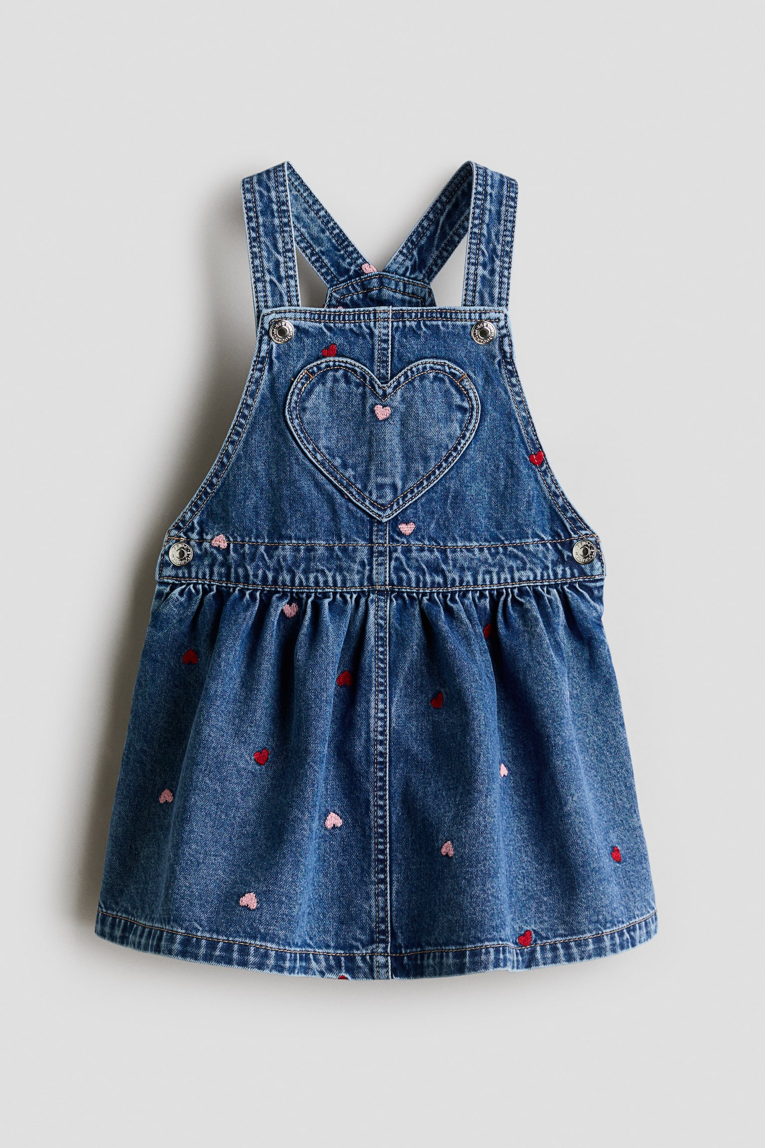 A blue denim pinafore dress is shown from the front, featuring a heart-shaped patch pocket on the bib and scattered embroidered pink and red hearts across the garment. The dress is designed with wide shoulder straps secured by metal buttons, side button closures at the waist, and a gathered, flared skirt.