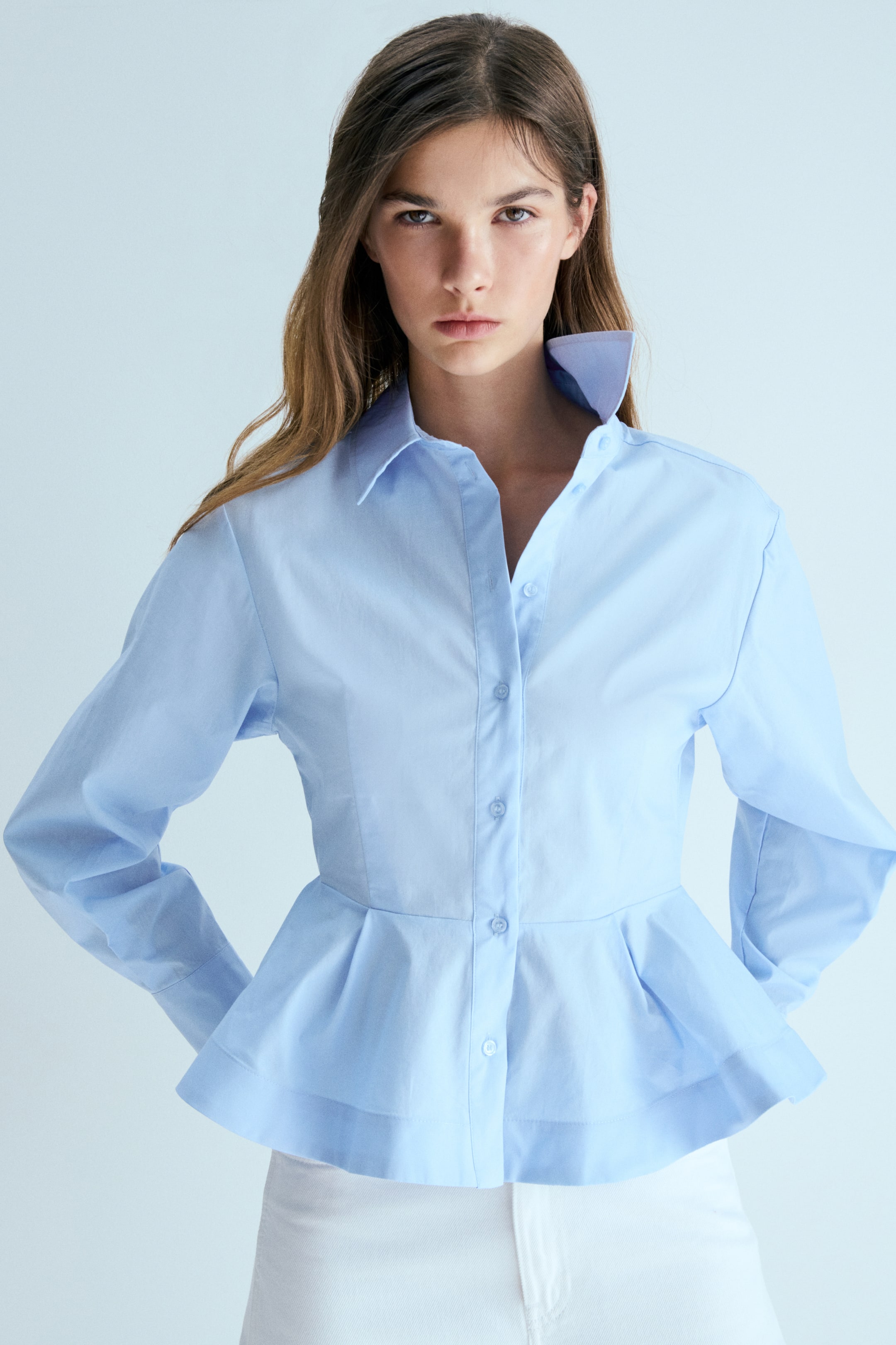 View larger image: A light blue peplum shirt is being worn, showcasing a full button-front placket and long sleeves with buttoned cuffs. Its pointed collar is partially standing up, and the shirt flares from a fitted waistline into a pleated peplum hem.