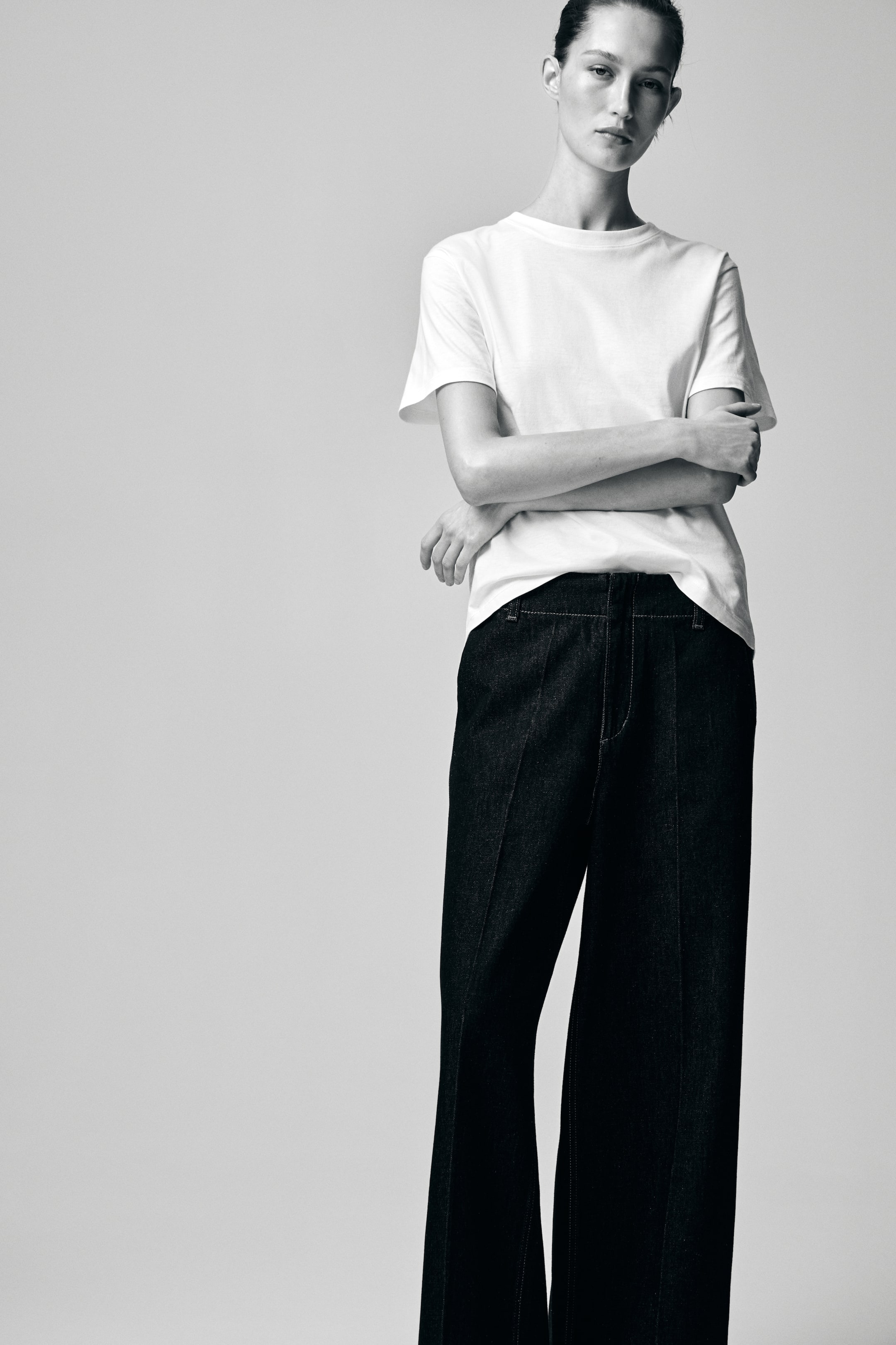 View larger image: A white crew-neck T-shirt with short sleeves and a relaxed fit is worn partially tucked into dark, high-waisted, wide-leg pants. Both items are presented from a front-facing view, with the pants showing visible topstitching along the front of each leg.