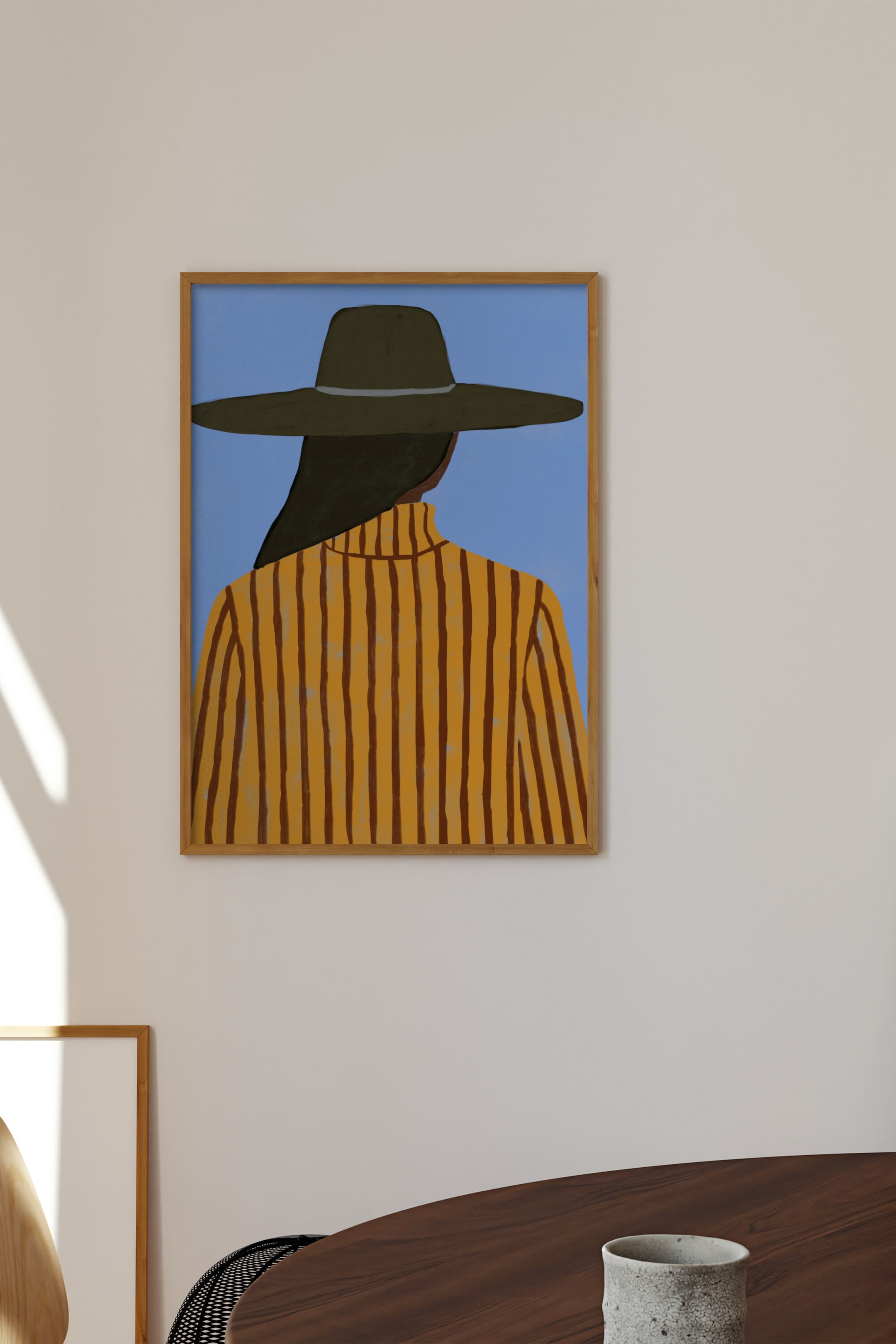 Ver imagem maior: Uma obra de arte emoldurada retrata a parte de trás de uma pessoa a usar um chapéu de aba larga verde-oliva escuro com uma faixa clara e uma camisola de gola alta amarela mostarda com riscas verticais castanho-escuras, tudo contra um fundo azul-claro, e está envolvida numa moldura de madeira natural.