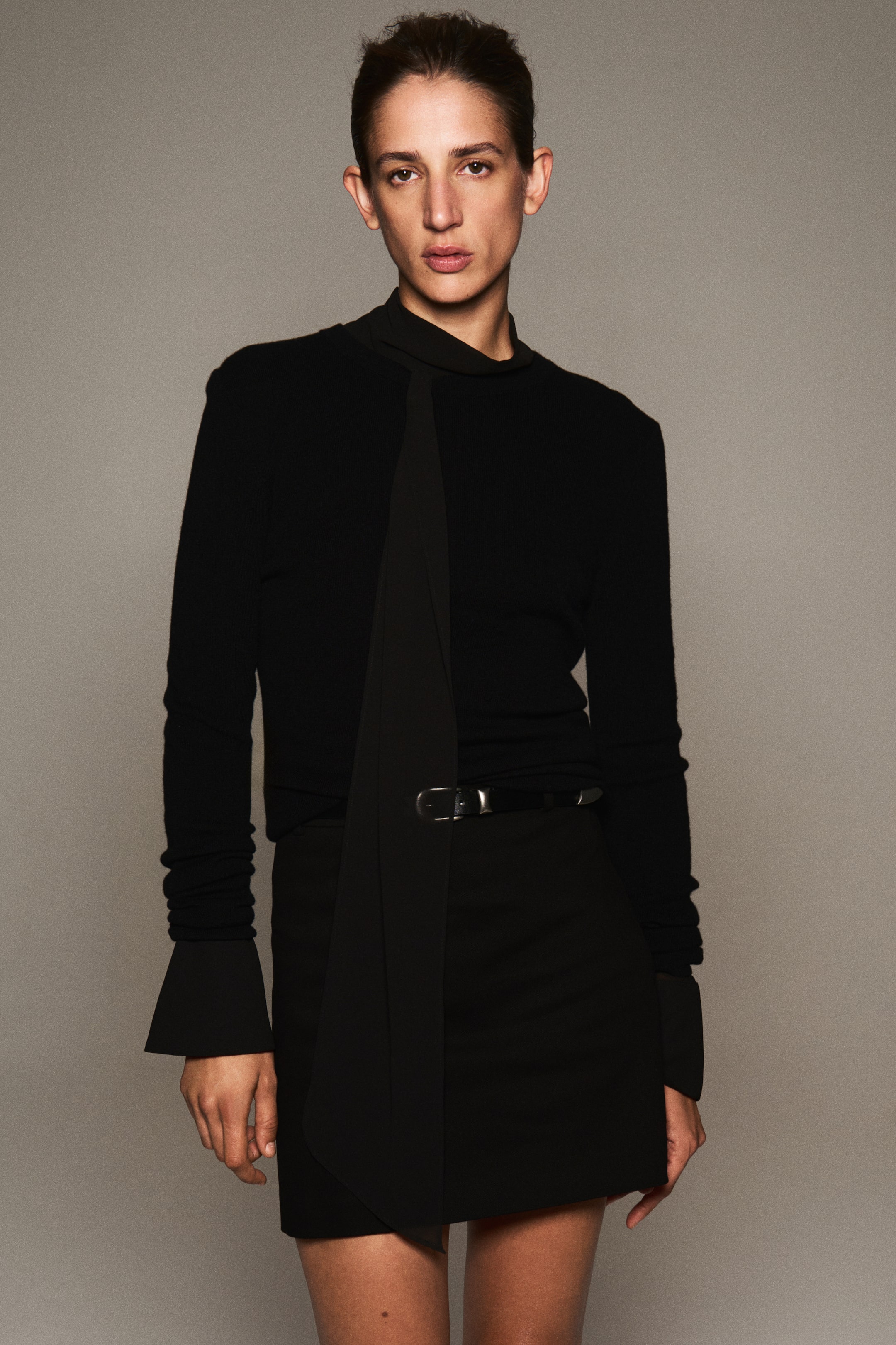 Agrandir l'image: Une mini-jupe noire ajustée à taille haute, dotée d’une fine ceinture réglable avec boucle argentée, est portée par le mannequin. La jupe est associée à un haut noir à manches longues, orné d’un long lien en tissu tombant sur le devant.