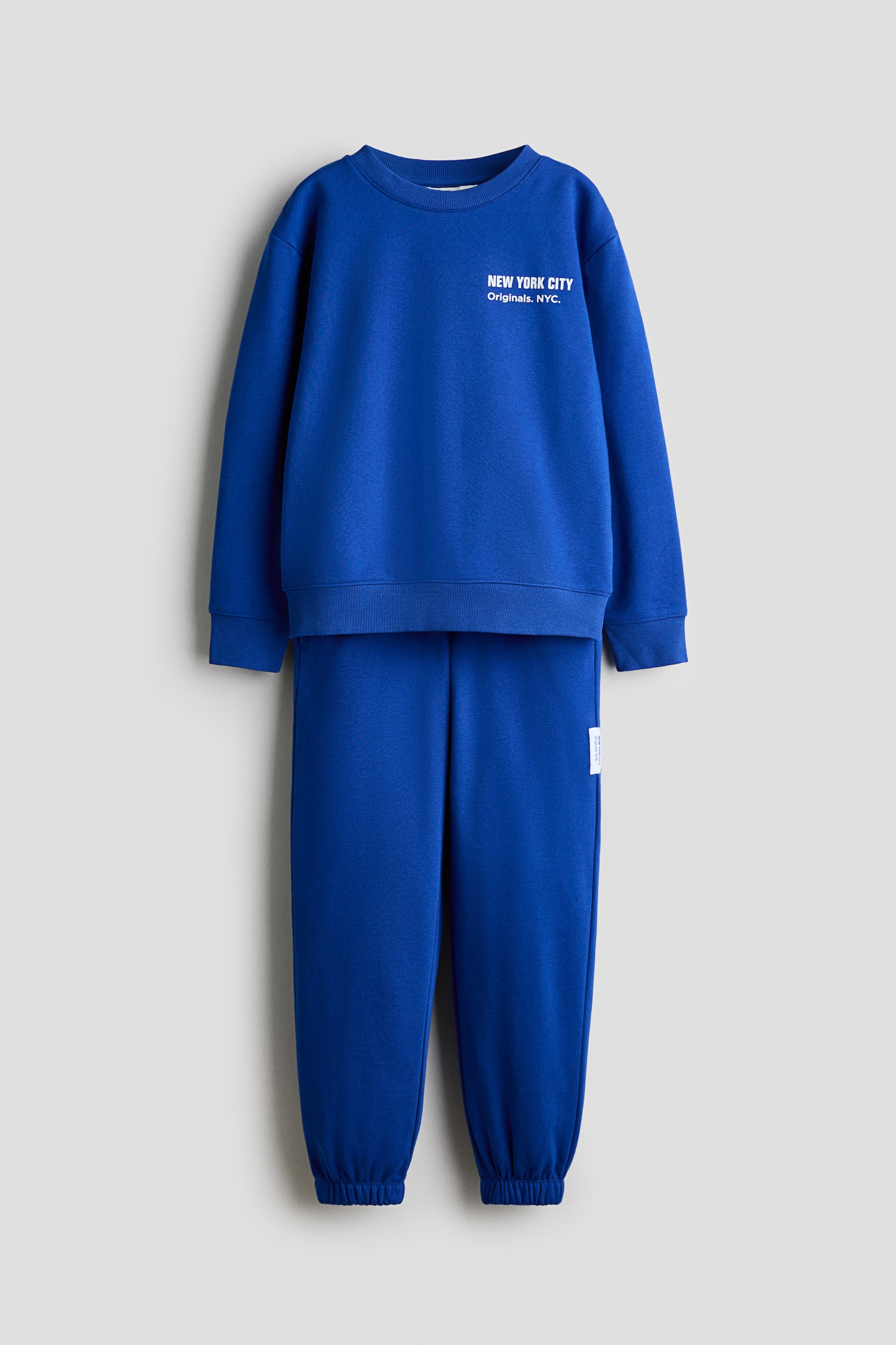 Dieses blaue zweiteilige Set besteht aus einem Sweatshirt und einer passenden Jogginghose, die von vorne gezeigt werden. Das Sweatshirt verfÃ¼gt Ã¼ber einen Rundhalsausschnitt, gerippte BÃ¼ndchen und einen gerippten Saum sowie einen kleinen weissen Aufdruck auf der linken Brustseite, wÃ¤hrend die Jogginghose elastische BÃ¼ndchen aufweist.