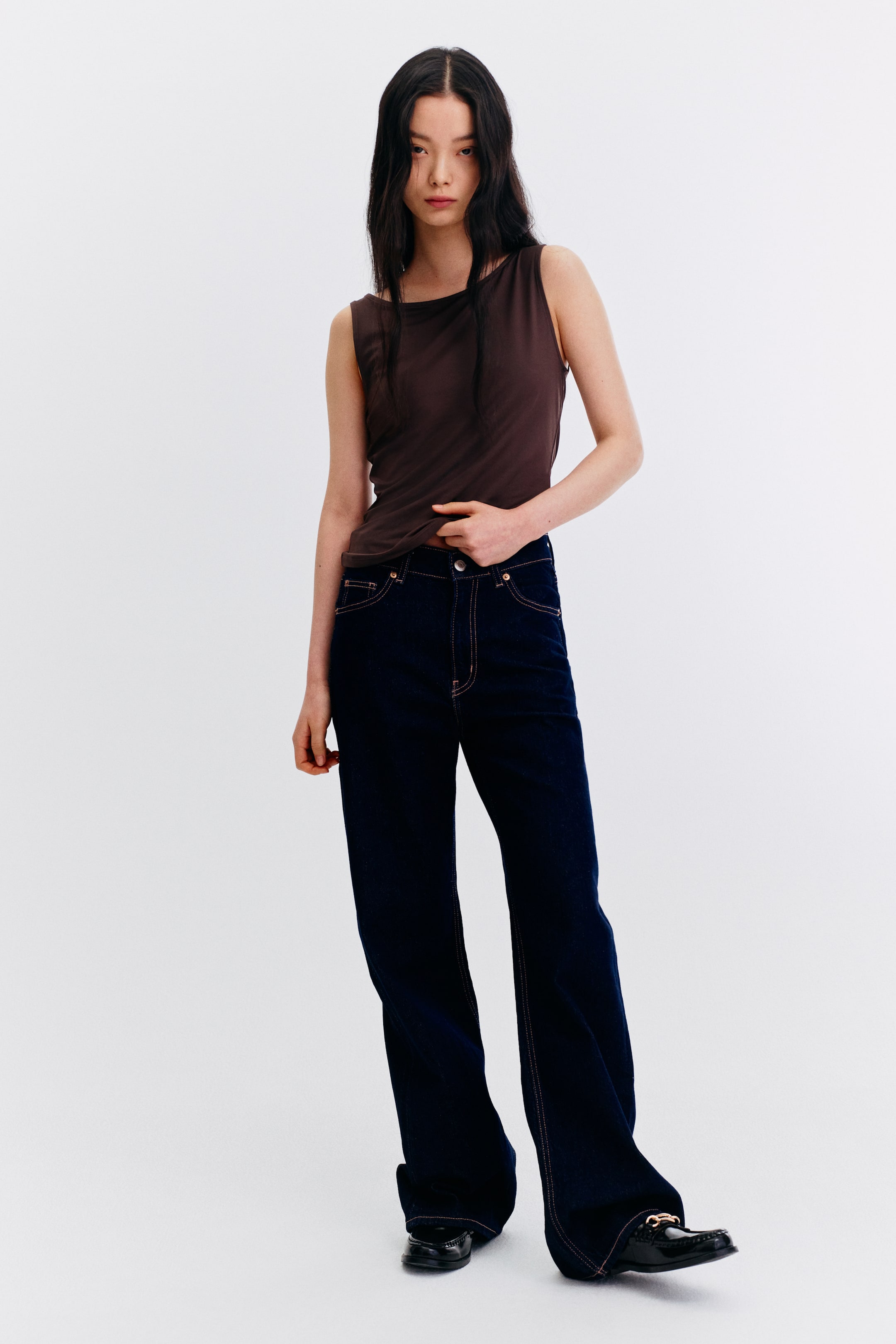 View larger image: The model dons a brown sleeveless top featuring a round neckline and a tailored fit. Paired with dark wash, wide-leg jeans and black loafers adorned with gold hardware, she gently places her hand on the waistband of the jeans.