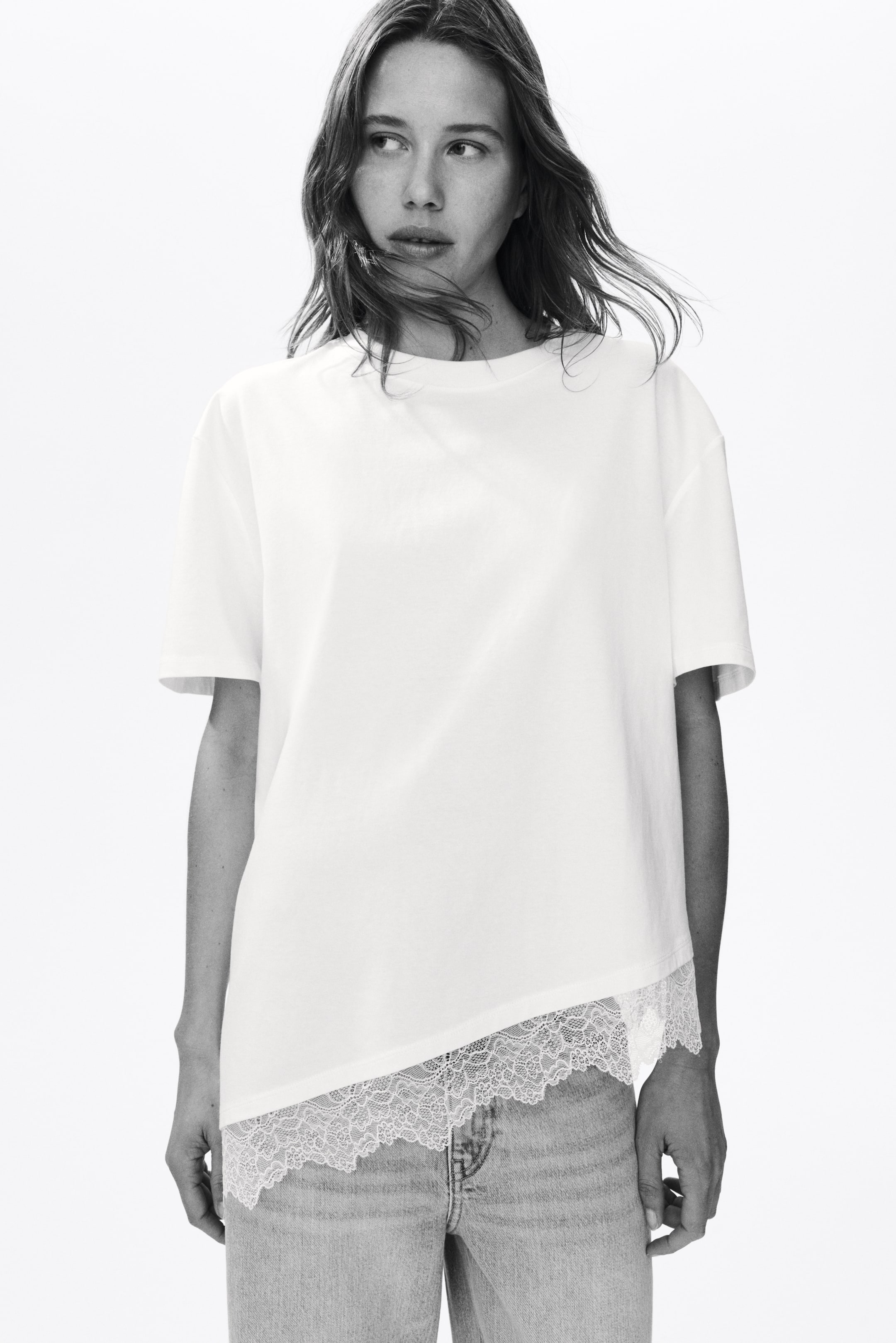 Grotere afbeelding bekijken: Een wit T-shirt heeft een ontspannen pasvorm met een ronde hals en korte, verlaagde mouwen. De asymmetrische zoom is voorzien van een witte kanten rand die over lichtgewassen jeans valt.