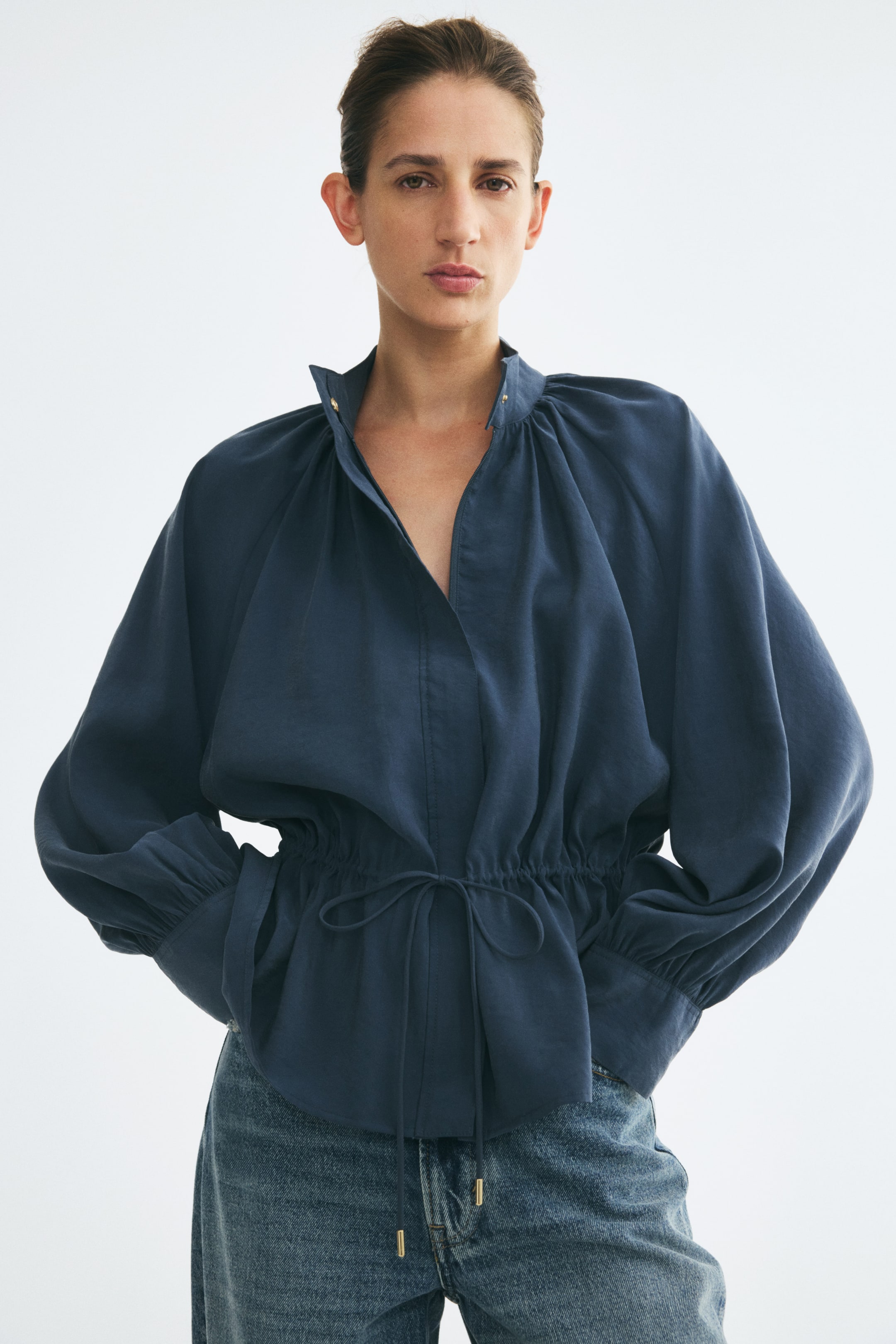 View larger image: A dark blue jacket featuring a stand collar, cuffed balloon sleeves, and a drawstring waist is worn by the model. The jacket has a flared peplum hem and is styled with a pair of blue denim jeans.