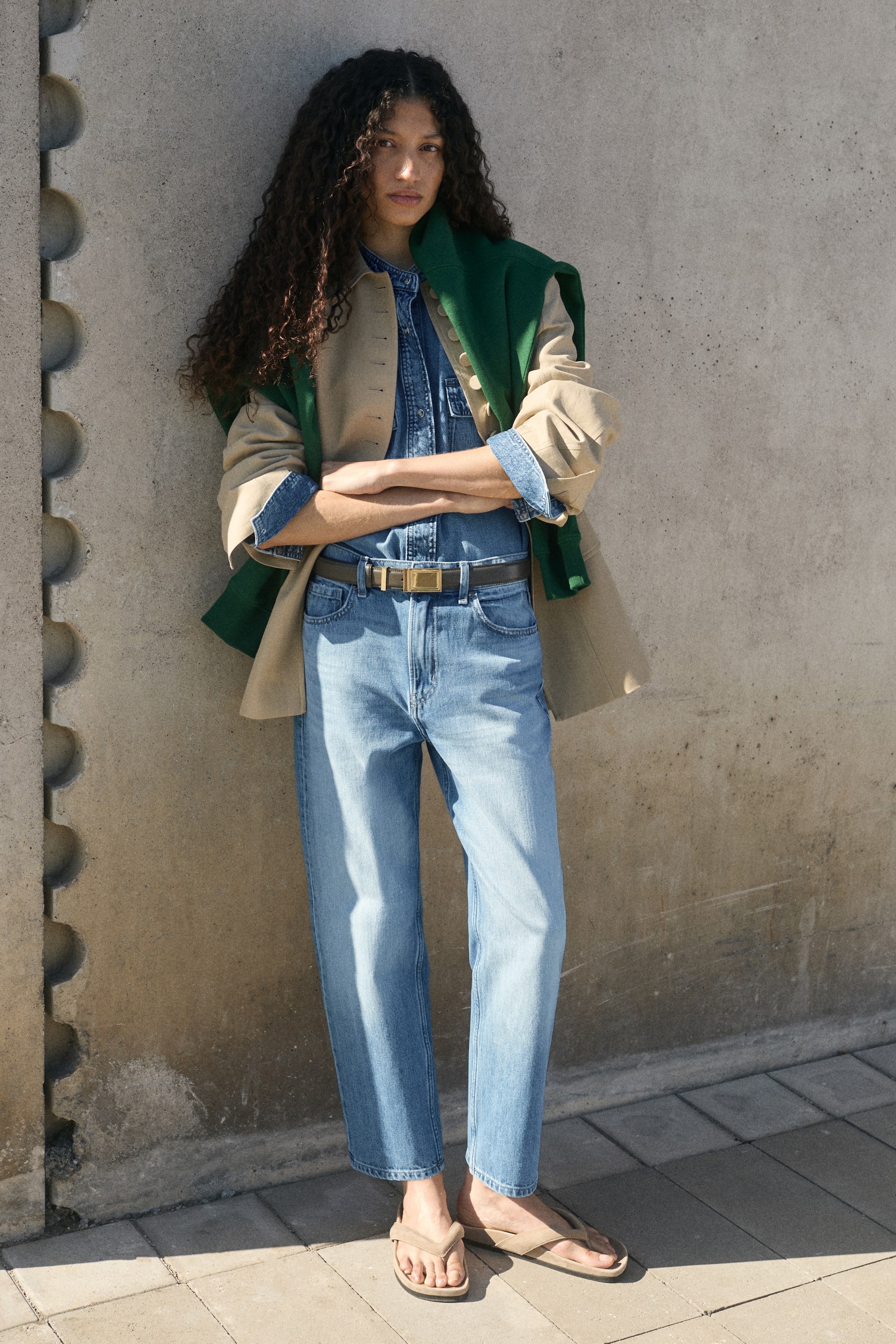 Visualizza l'immagine ingrandita: Il modello indossa jeans in denim dal lavaggio azzurro chiaro, a gamba dritta, con vestibilità regular e orlo alla caviglia. I jeans sono abbinati a una camicia in denim coordinata infilata dentro, stretta da una cintura marrone, a una giacca beige aperta, a un maglione verde drappeggiato sulle spalle e a sandali flat con fascia infradito.