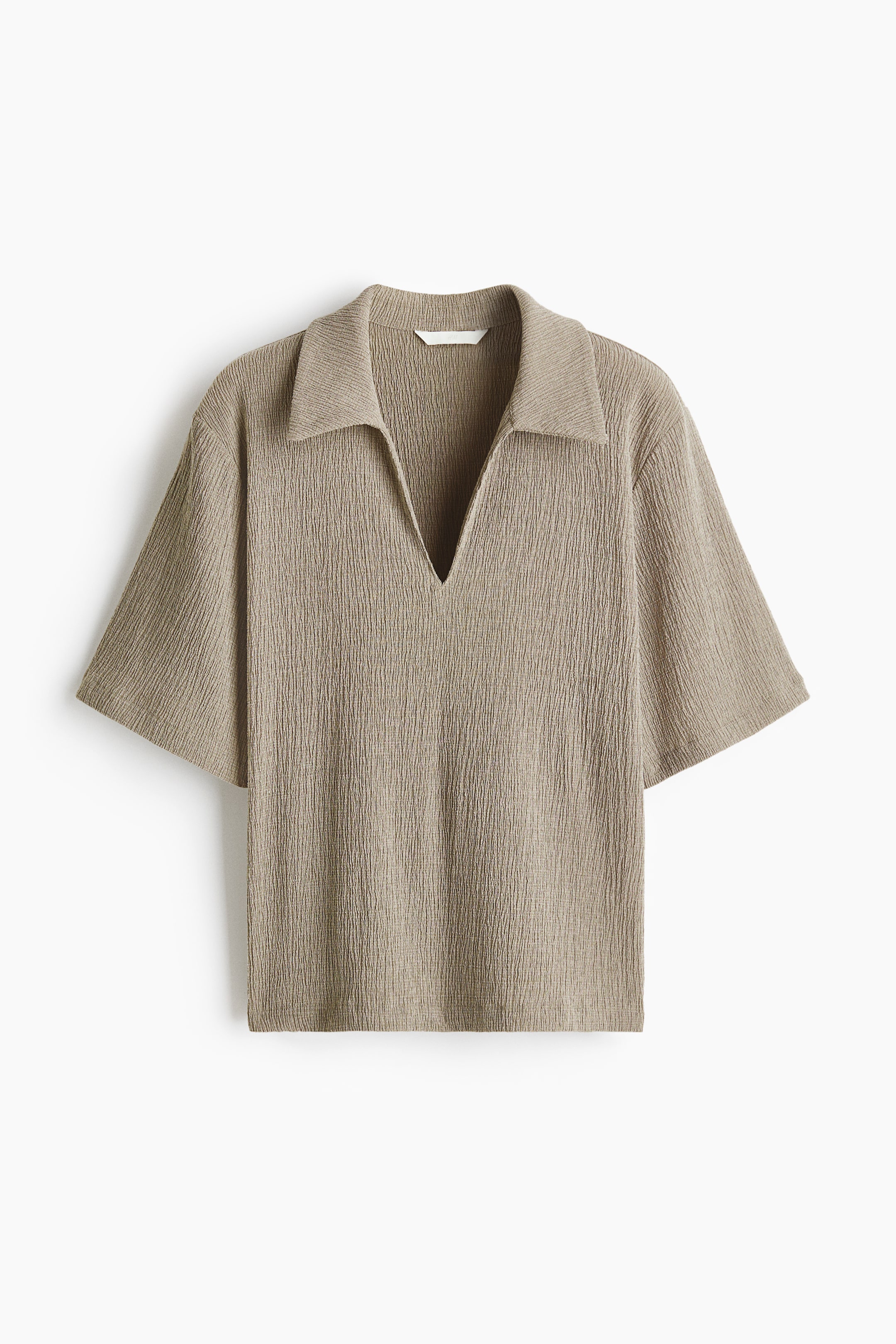 Visa större bild: En löst sittande polotröja i ljusbrun skrynklad jersey visas framifrån, med en öppen krage som bildar en V-ringning, nedhasade axlar och korta ärmar.
