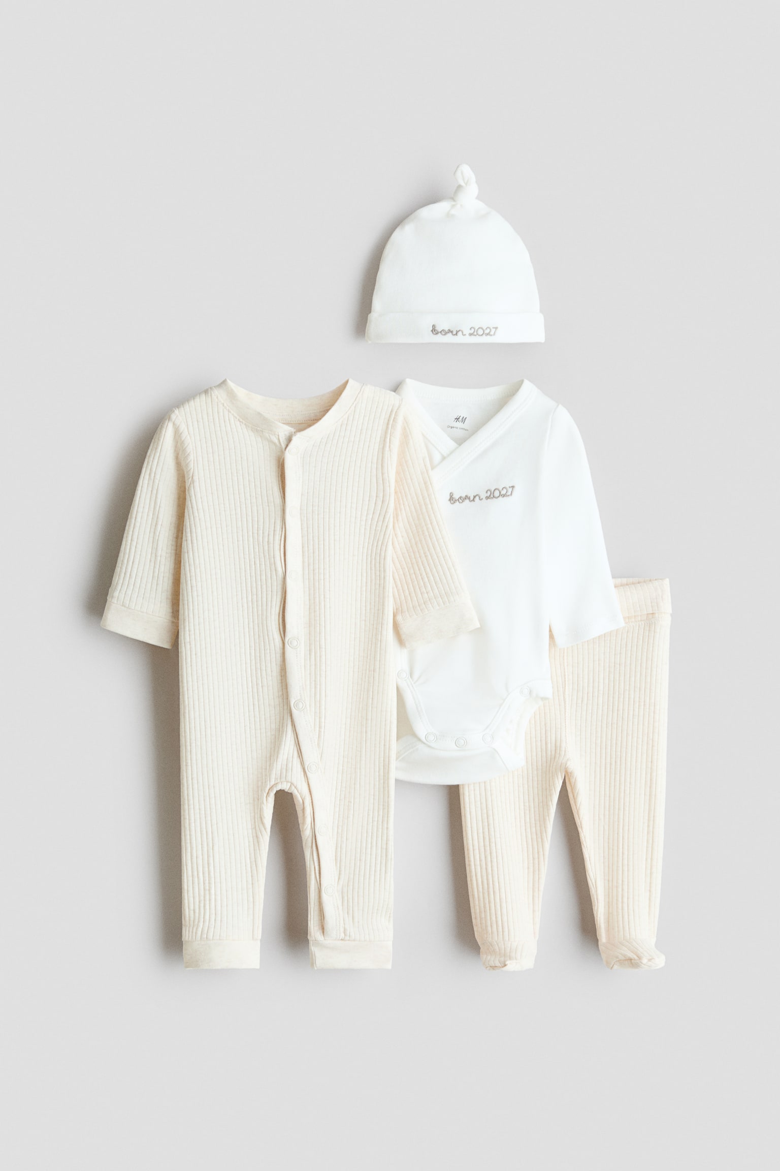 Een katoenen jersey cadeauset uit 4 delen presenteert een crèmekleurige, geribde jumpsuit met lange mouwen en drukknoopsluitingen aan de voorzijde, samen met een witte wrap-over body met lange mouwen en "born 2027"-borduursel op de borst. De set toont ook bijpassende crèmekleurige geribde broek met voetjes en een witte knoopmuts met bijpassend borduursel op de omgeslagen boord.