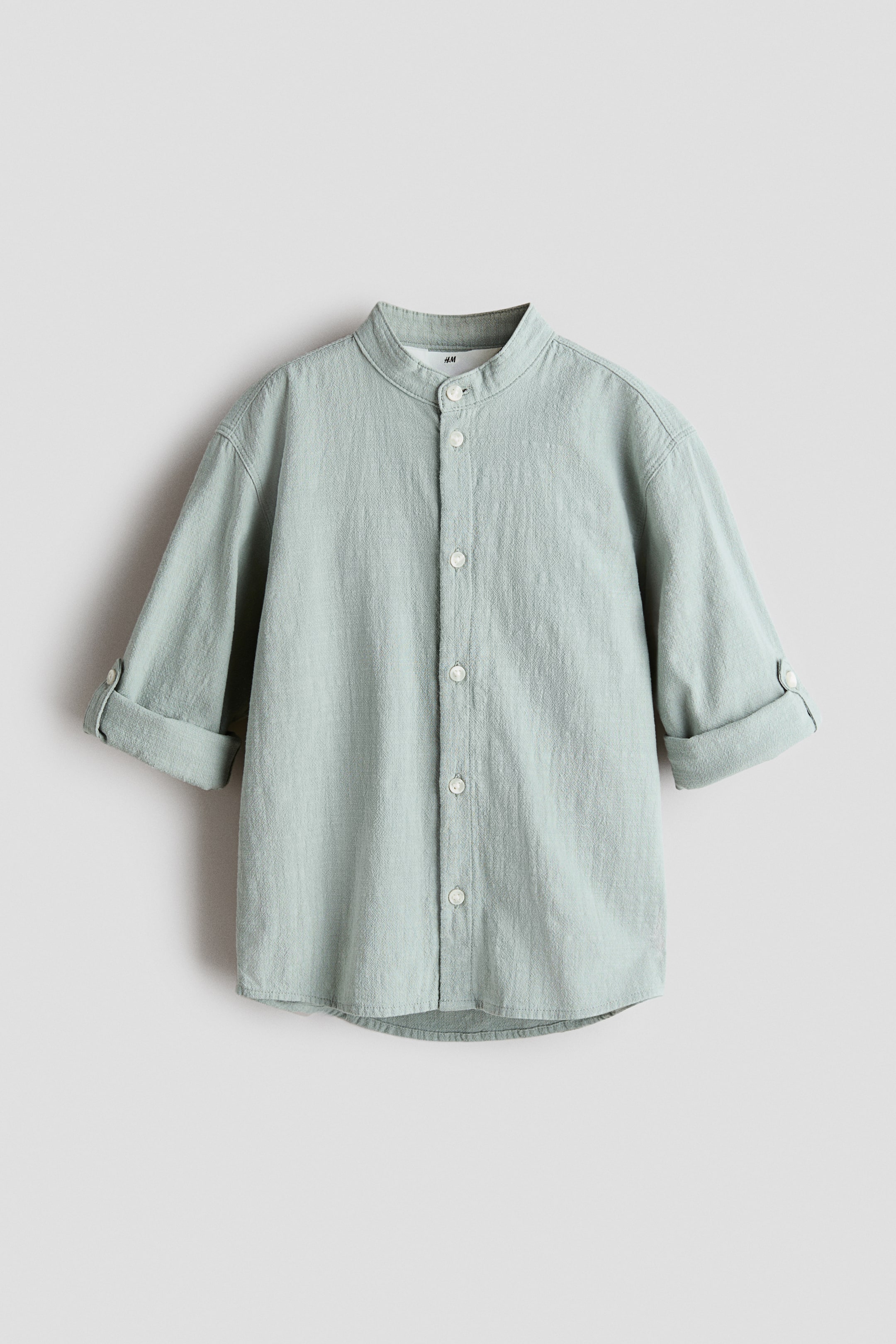View larger image: A light green shirt in a soft, textured weave is shown from the front, featuring a band collar and white buttons down the front. The long sleeves are rolled up and secured with buttoned tabs, showcasing the relaxed style and lightweight fabric of the garment.