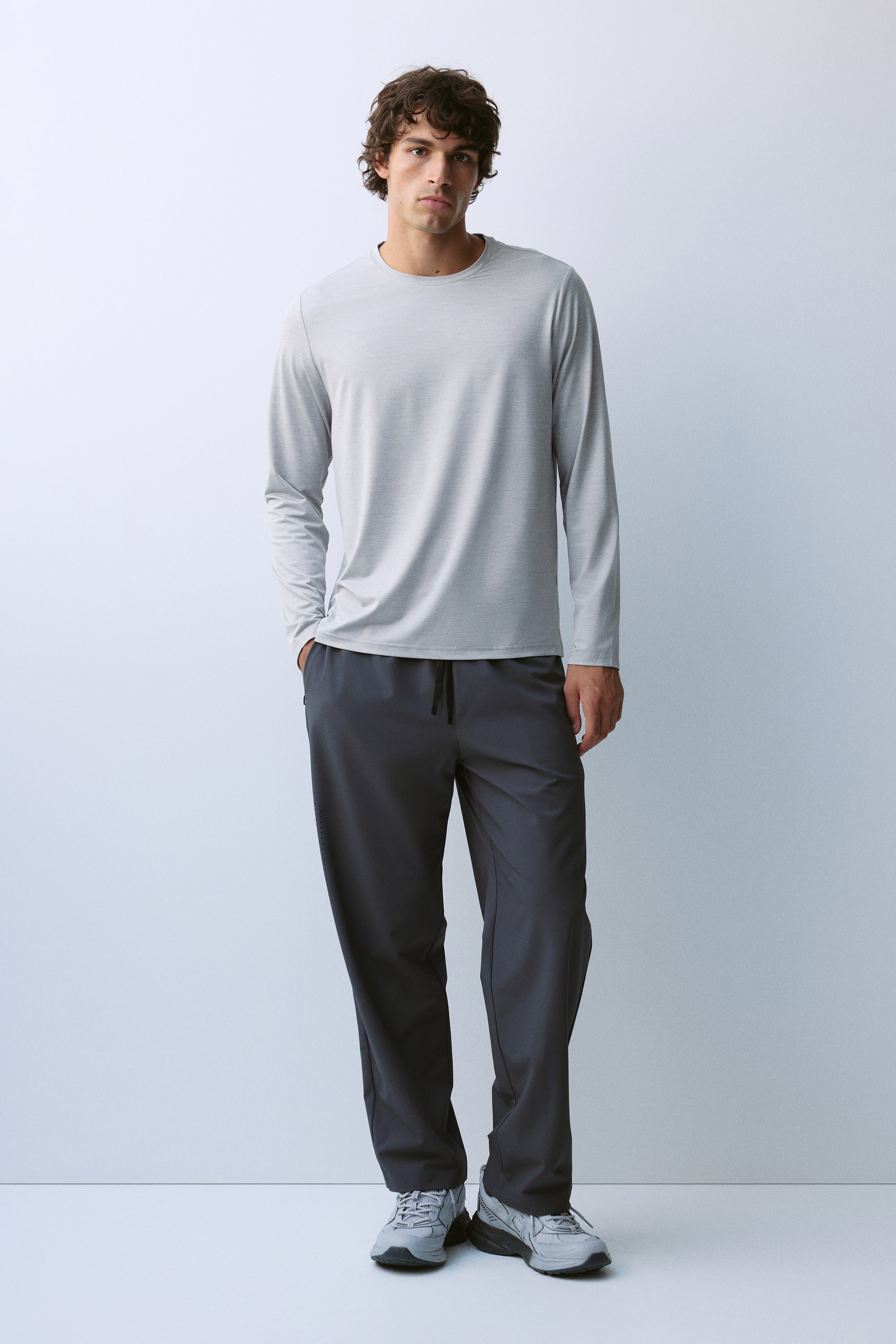 View larger image: The model is wearing a light grey long-sleeve crewneck T-shirt and dark grey drawstring trousers. He is standing with one hand in his trouser pocket, completing the look with grey athletic sneakers.
