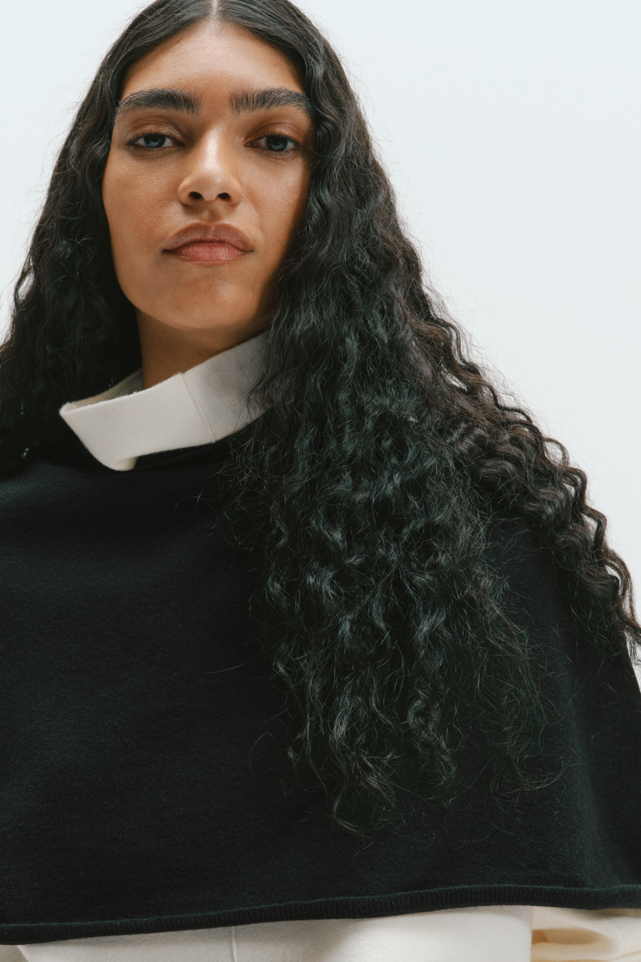 Agrandir l'image: Une cape noire en maille douce est portée par le mannequin, superposée à un vêtement crème à col montant. La cape tombe sur les épaules, mettant en valeur sa texture lisse.
