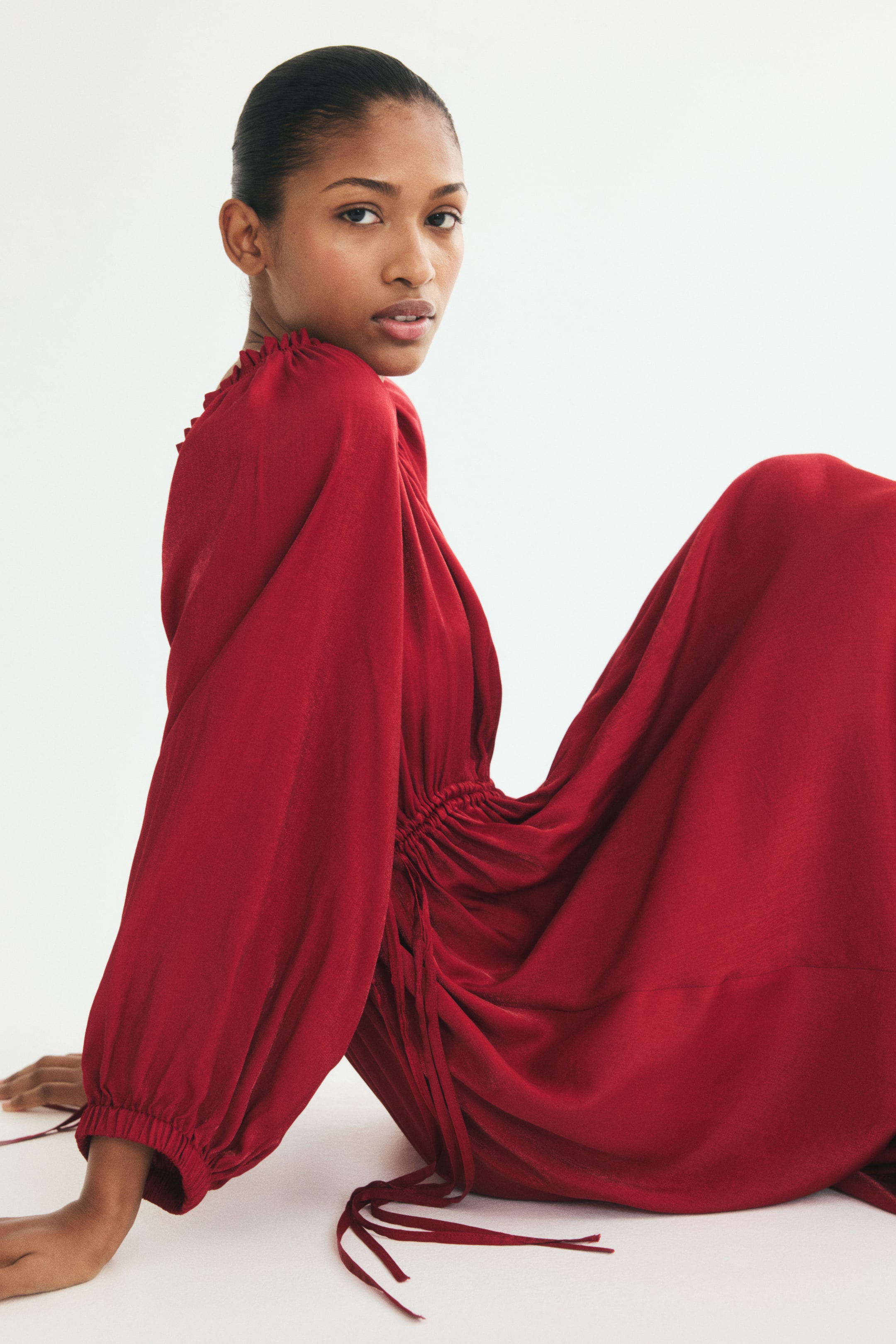 View larger image: A long, loose-fit red dress is shown from a side profile facing left, highlighting its draped, flowing silhouette. The dress features a high, ruffled neckline, long balloon sleeves with elasticated cuffs, and a gathered waist with long, hanging drawstrings.