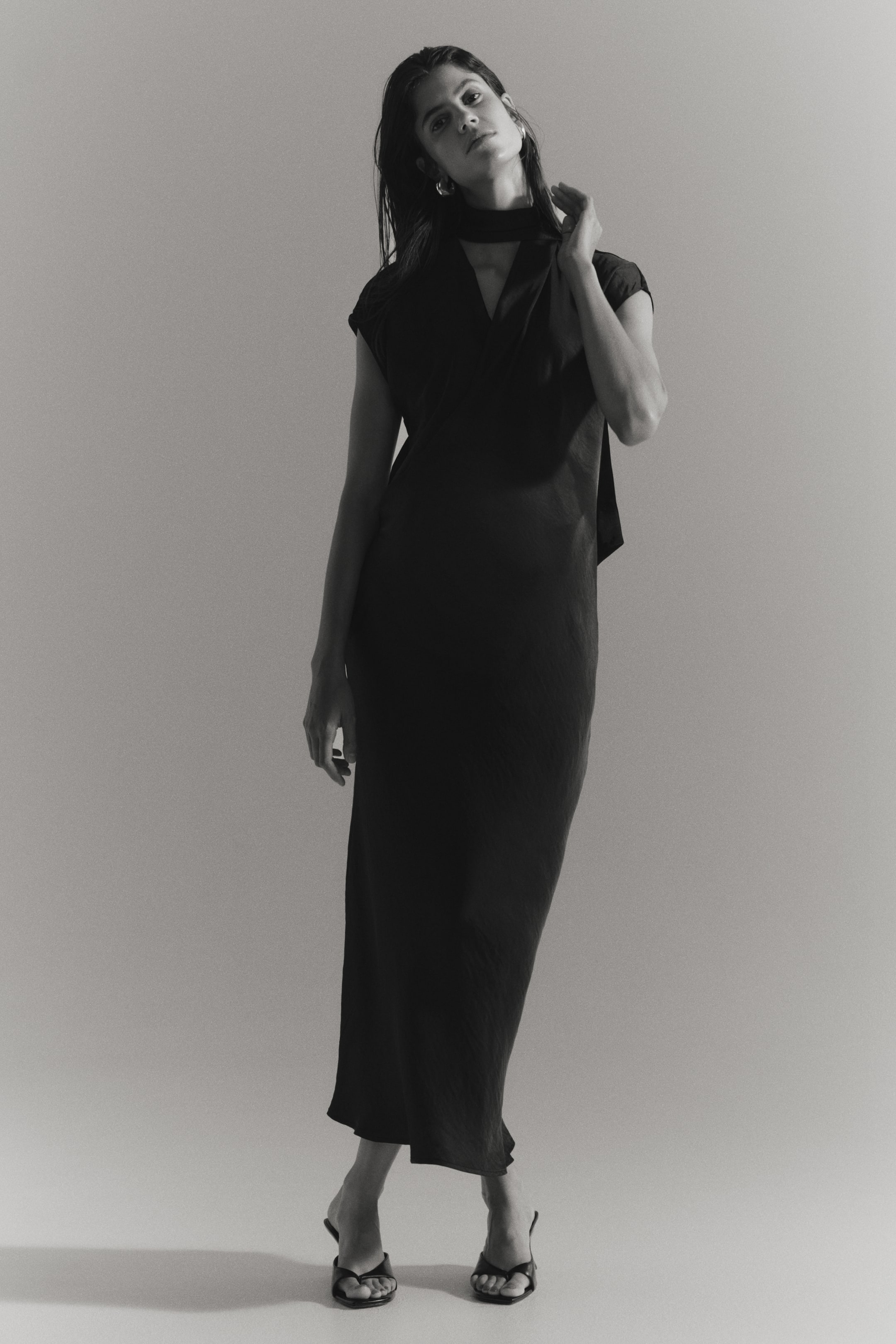 View larger image: A calf-length dark satin nursing dress is worn by the model, featuring a wrap-front design, cap sleeves, and a V-neckline. An attached long scarf detail is wrapped around the neck, which the model is lightly touching while standing in black heeled slide sandals.